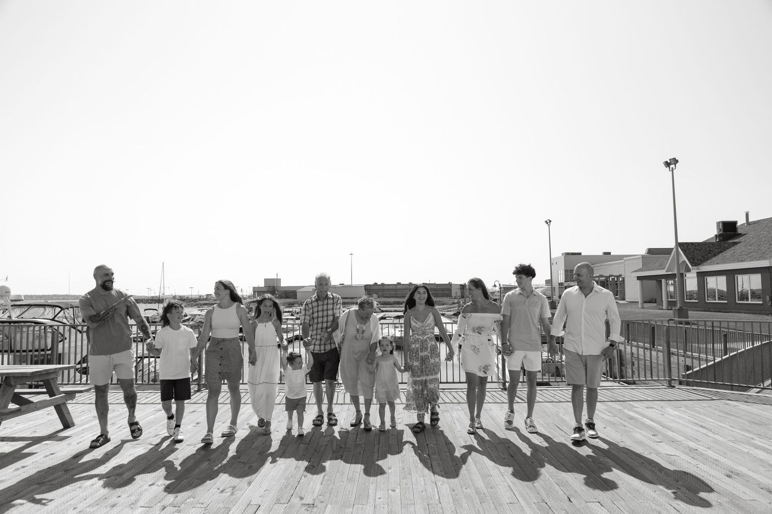 Black and white photo of a large group holding hands on a sunny boardwalk near waterfront buildings.