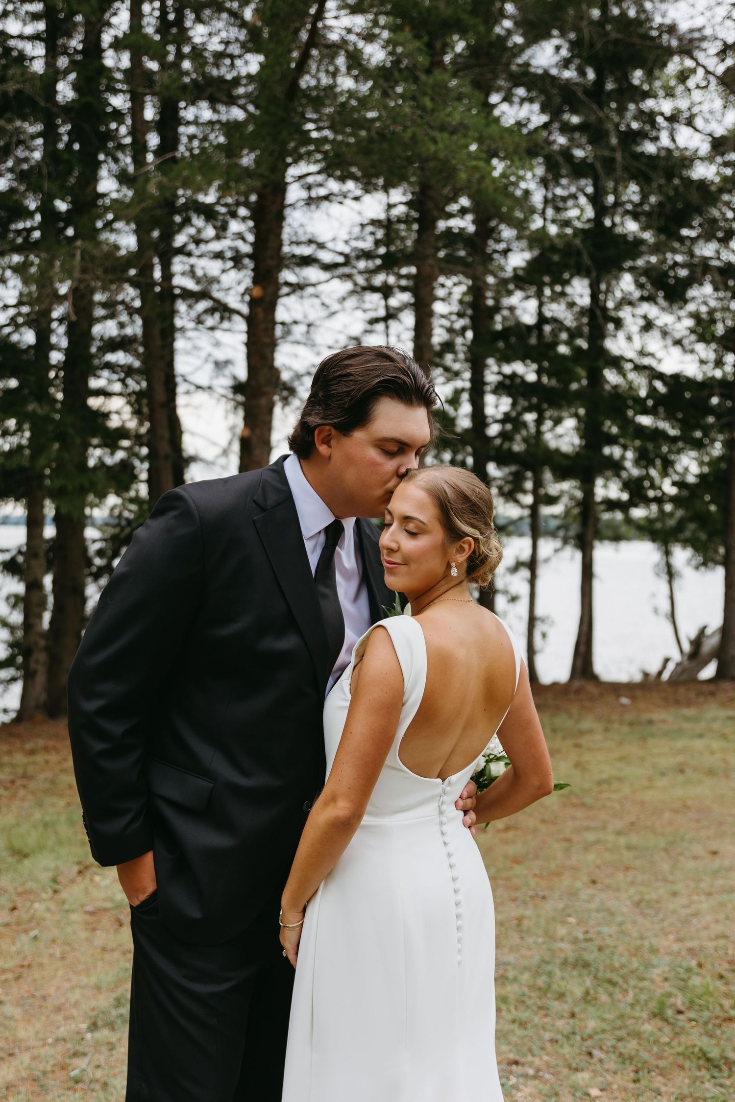 A romantic moment captured between newlyweds in an outdoor woodland setting with tall pine trees in the background.