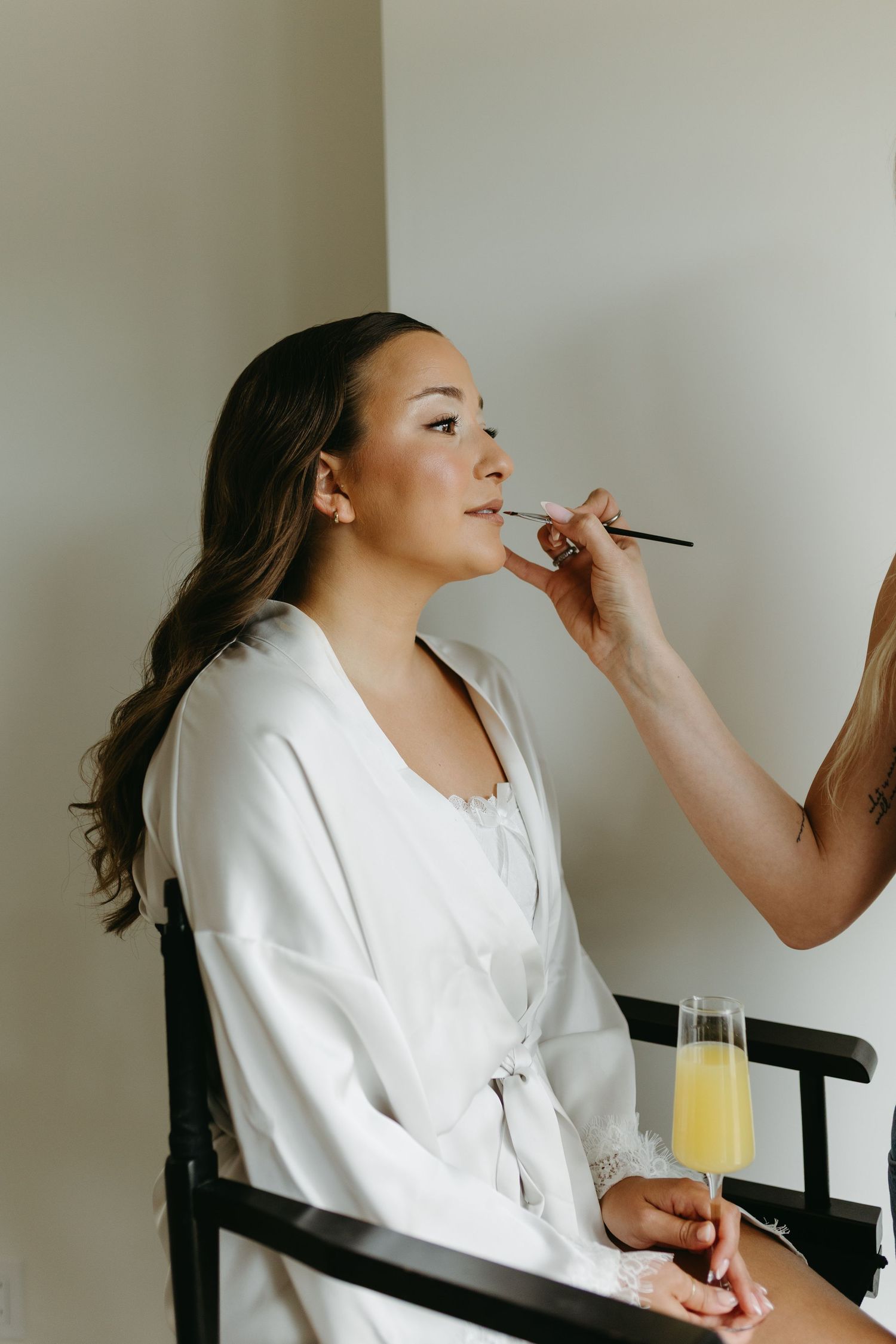 A person in a white robe gets makeup applied while holding a glass of orange juice during wedding preparations.