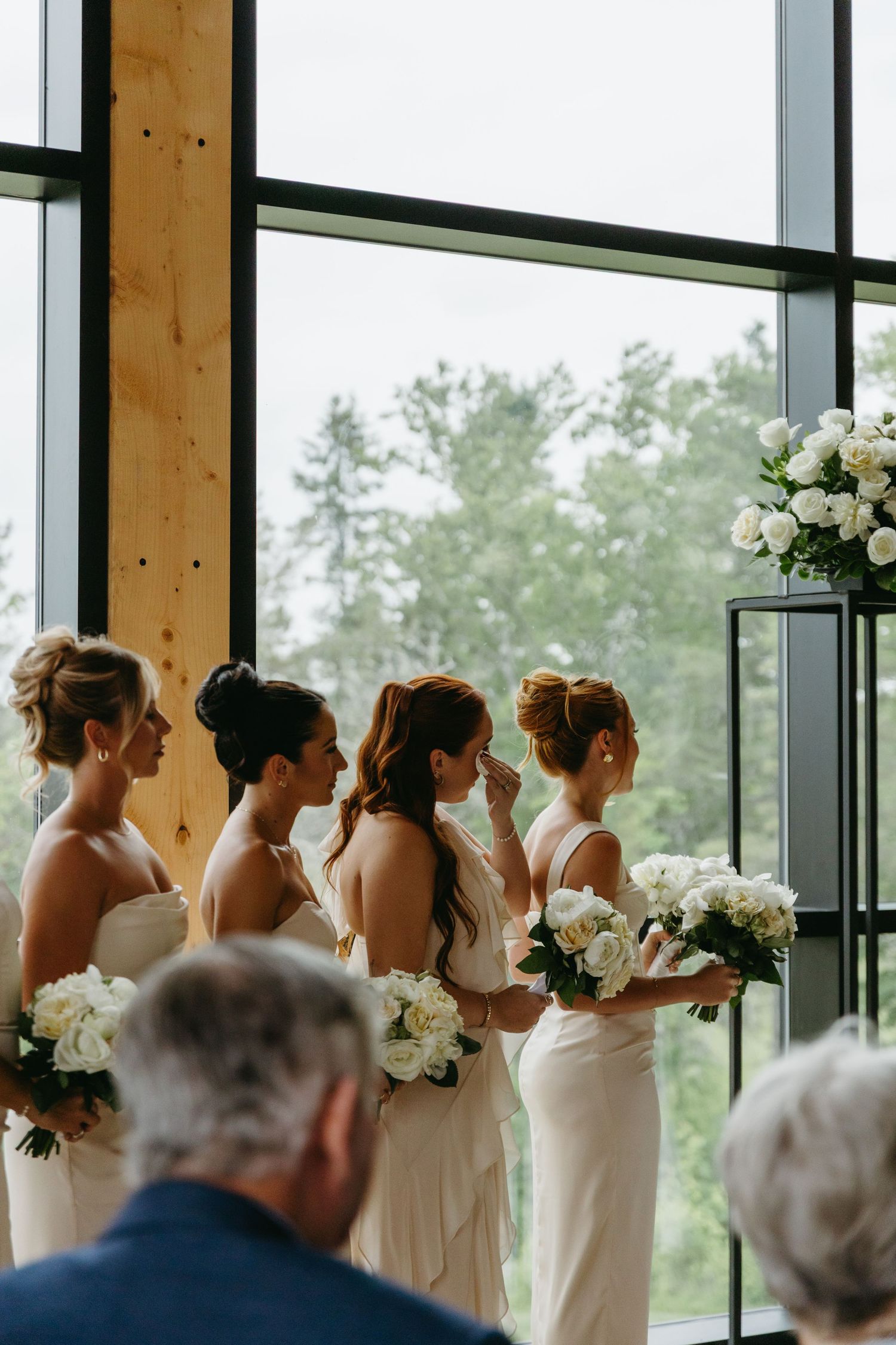 Bridesmaids in white dresses stand in line by large windows overlooking a scenic forest view.