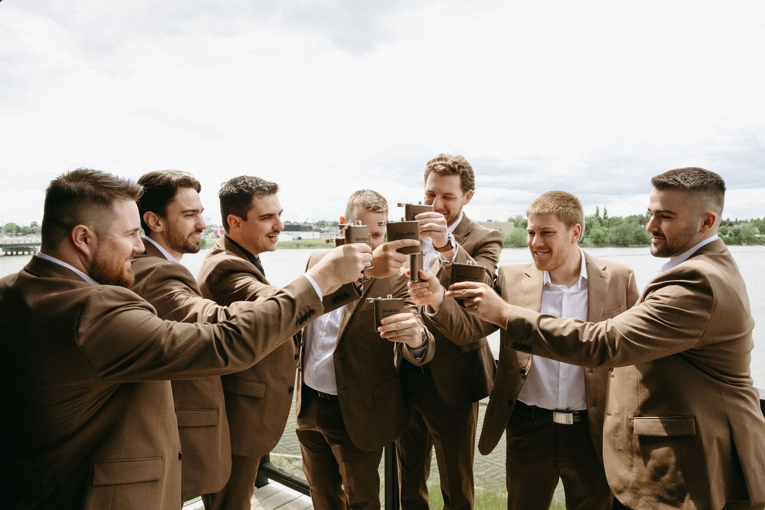 A group of groomsmen in brown suits raise their glasses in a celebratory toast outdoors.