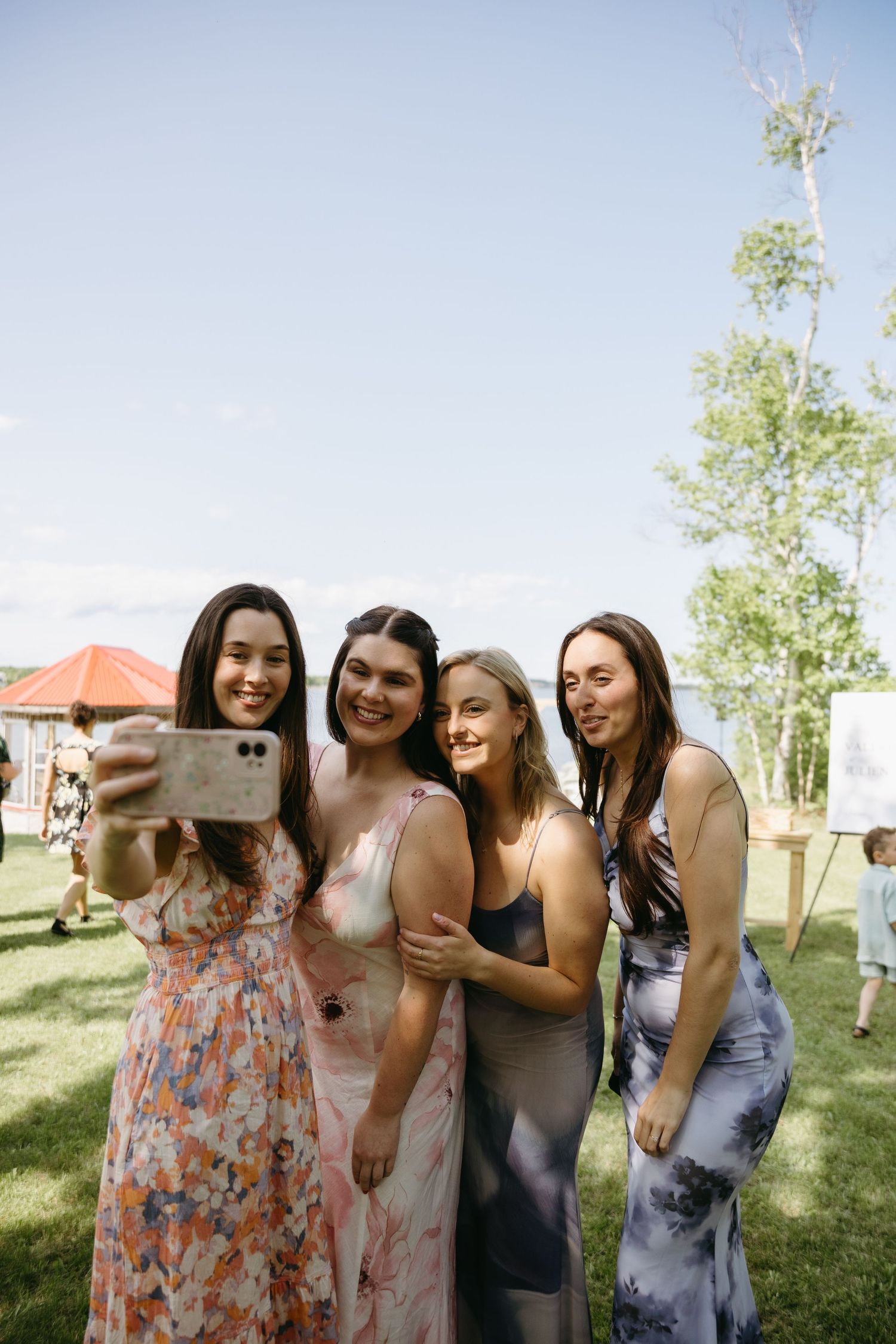 A group of friends in summer dresses take a selfie together on a sunny day in a grassy outdoor setting.