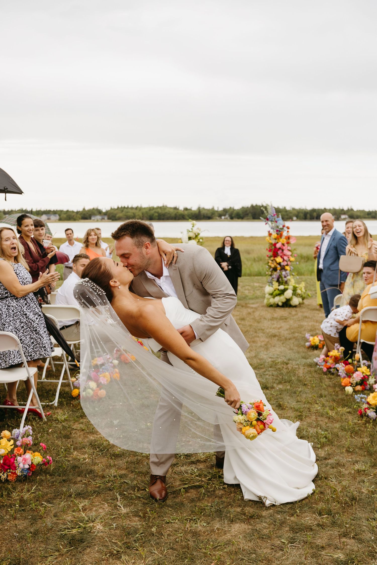 Newlyweds share romantic dip kiss during outdoor lakeside wedding ceremony with floral decorations and guests watching.