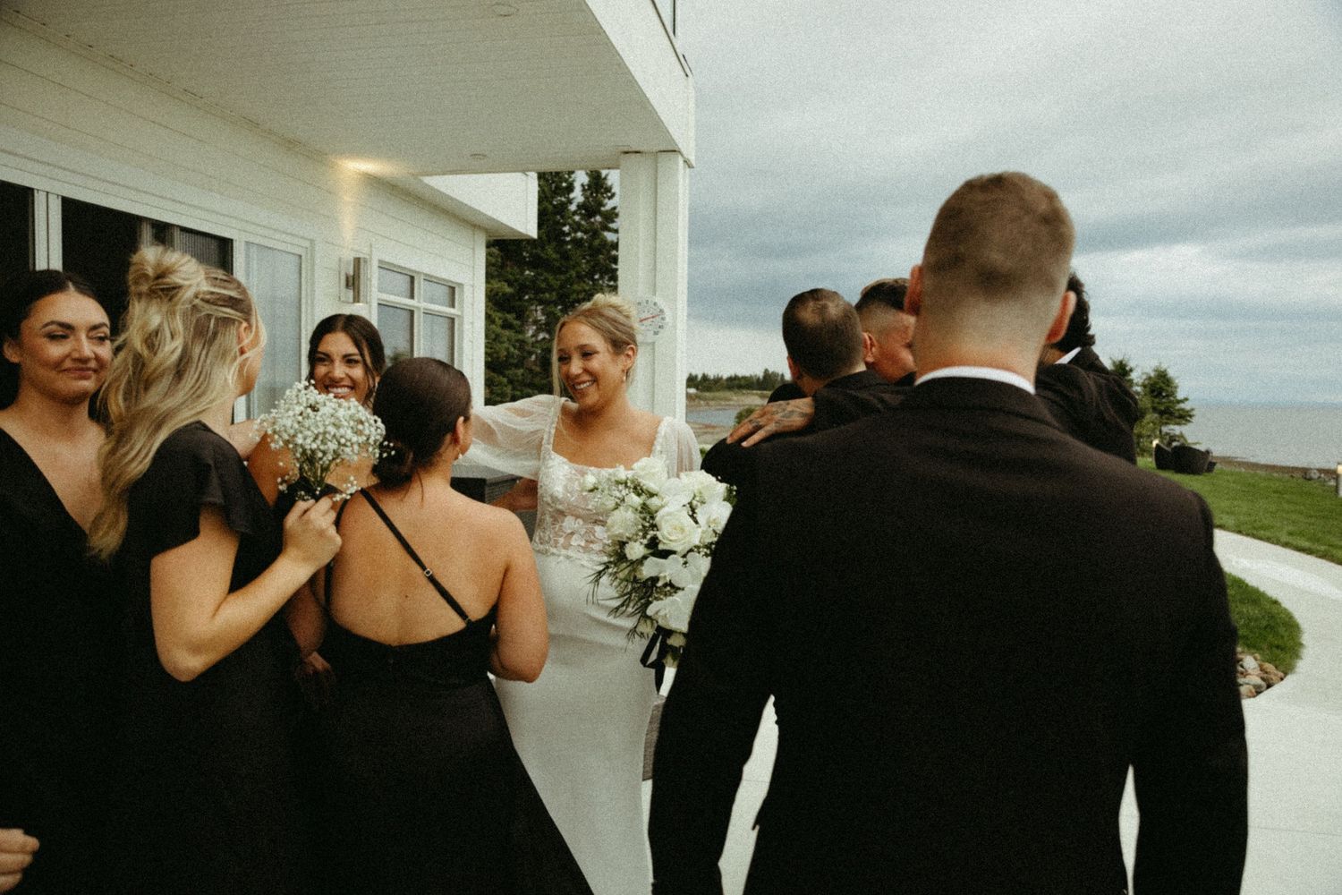 Wedding party celebrates together outside on a cloudy day with bridesmaids in black dresses and bride holding white bouquet.