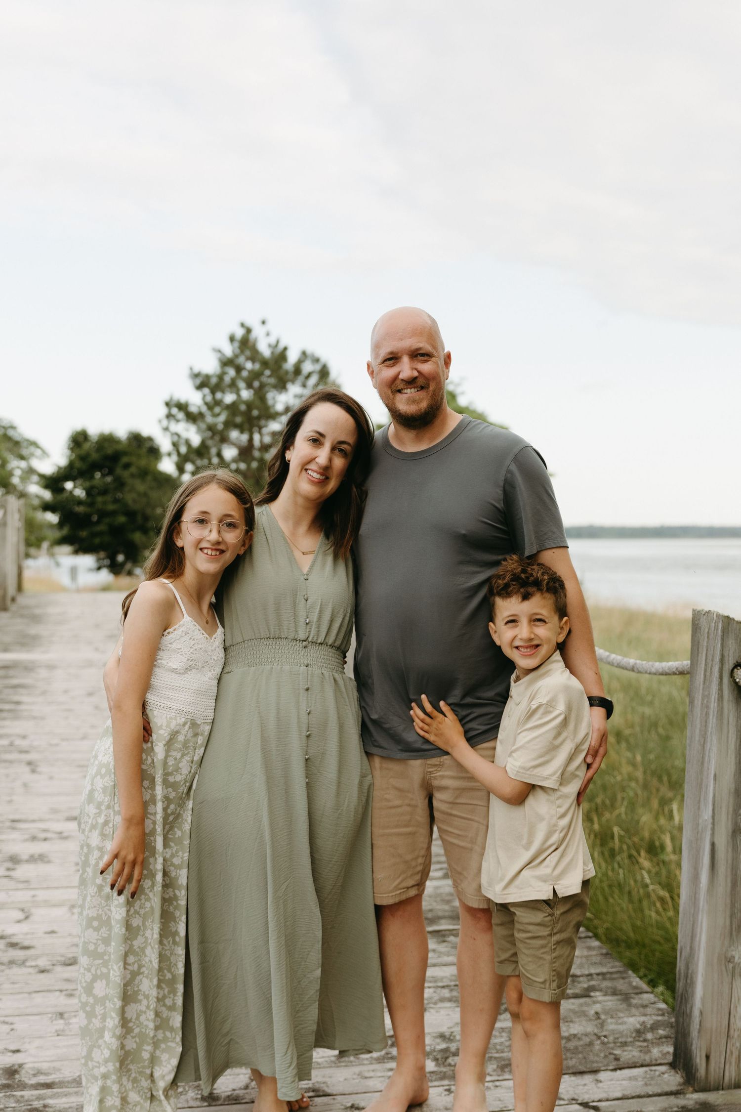 A family poses together on a wooden boardwalk by the beach in coordinating neutral outfits.