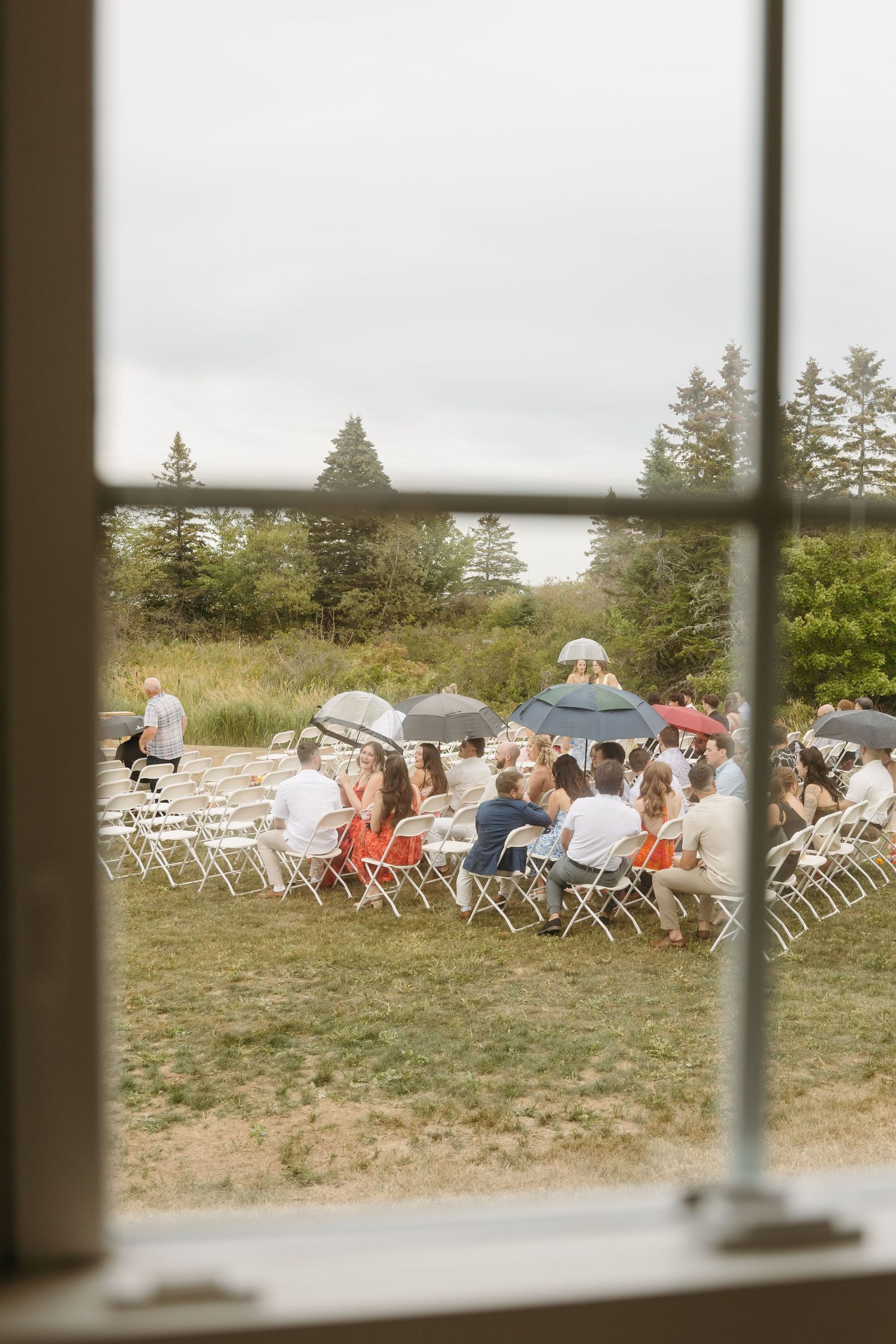 View through a window of outdoor wedding ceremony seating with white folding chairs and evergreen trees in background.