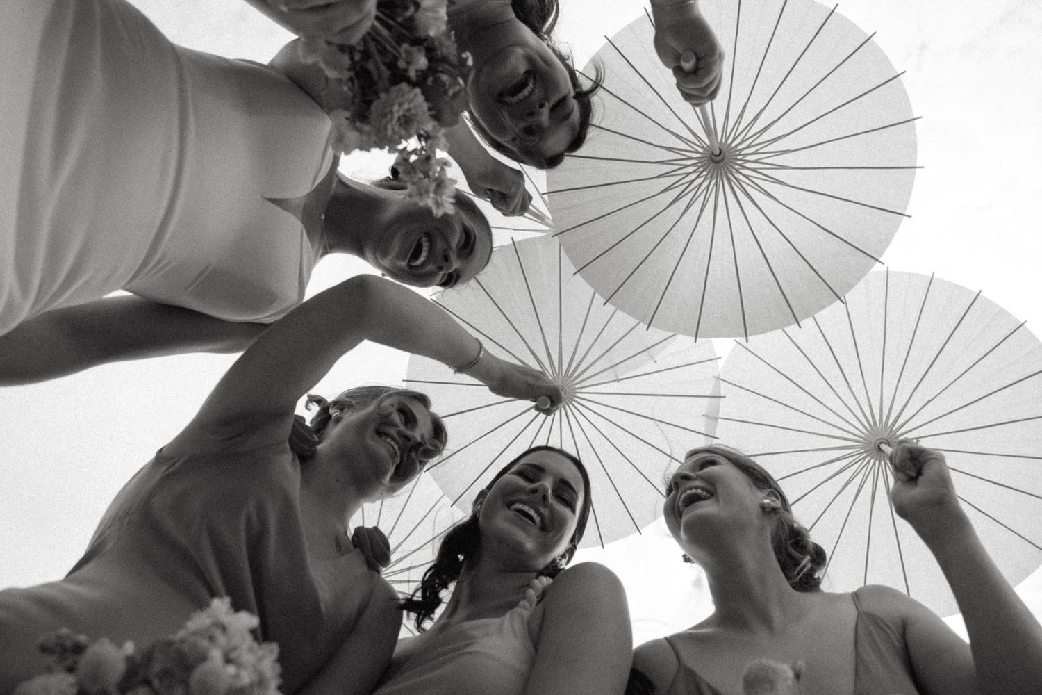 A joyful black and white photo showing a bride and bridesmaids with white parasols laughing together outdoors.
