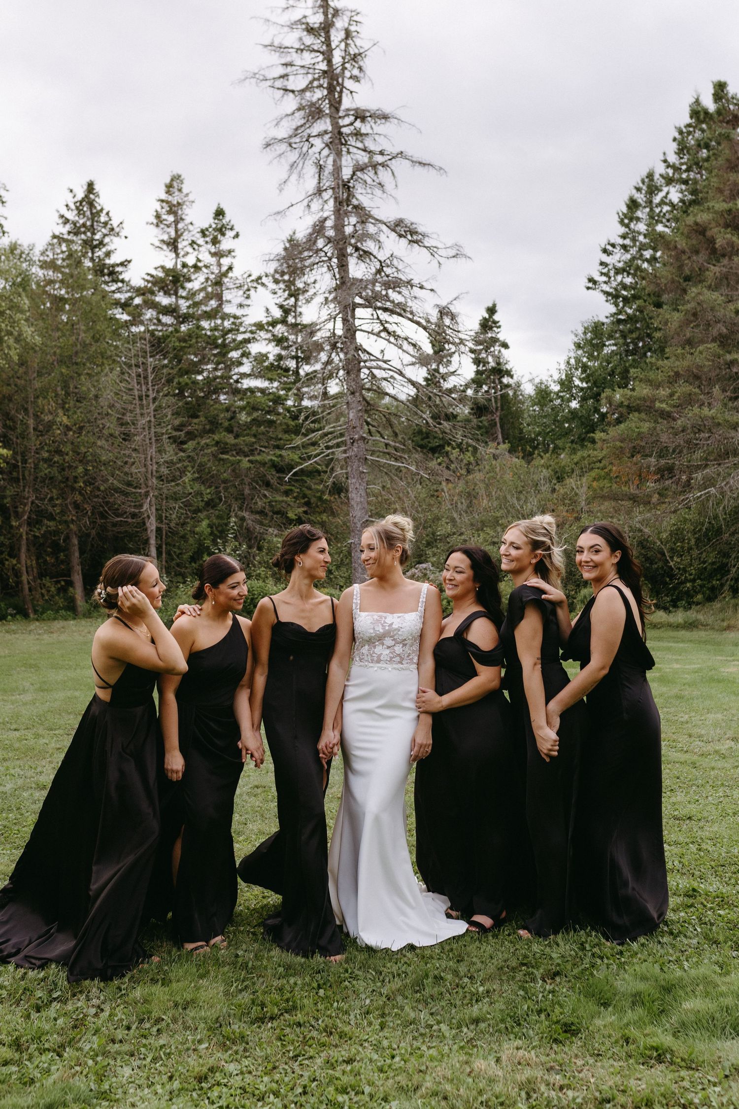 Wedding party in black dresses and white gown pose together on grassy field with evergreen trees in background.