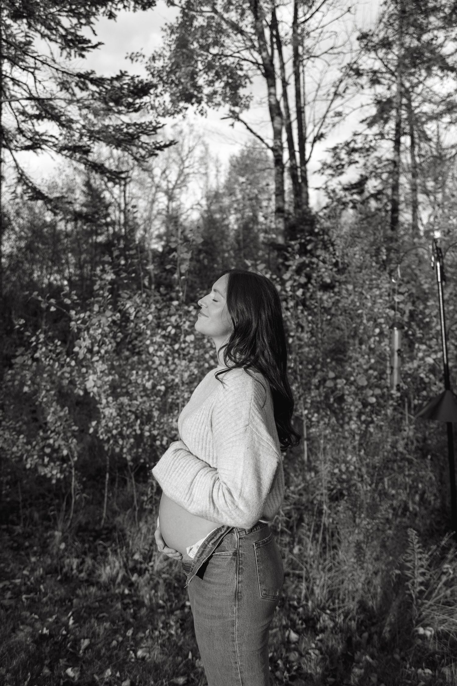 Black and white portrait of a person in a sweater and jeans standing in a forest setting, looking upward thoughtfully.