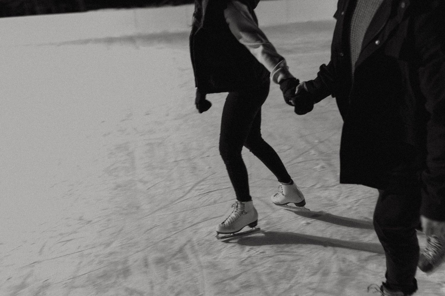 Black and white photo of two people holding hands while ice skating, with one person wearing sneakers on the ice.