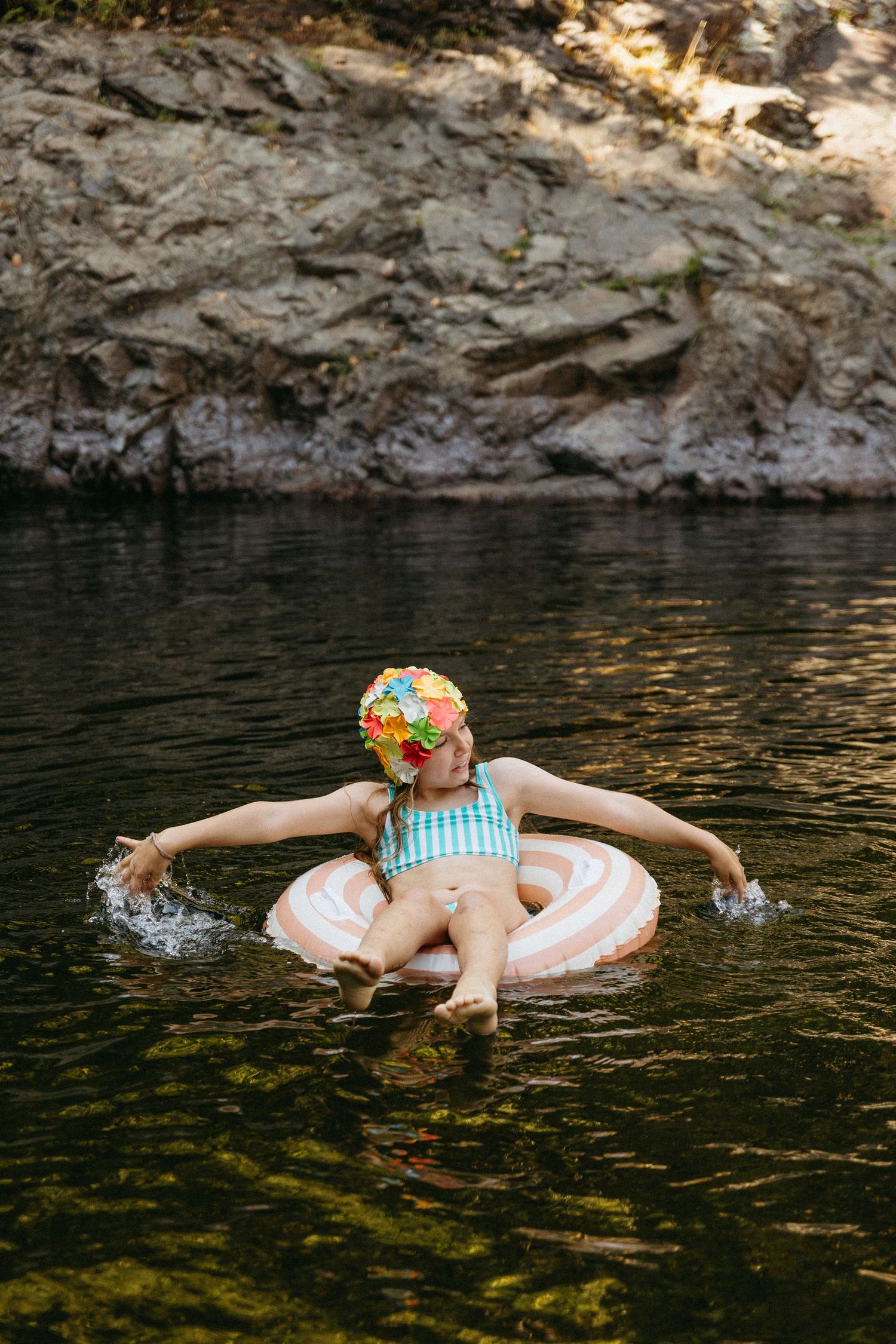 A person floats peacefully in a pink inner tube on dark water surrounded by rocky cliffs on a summer day.