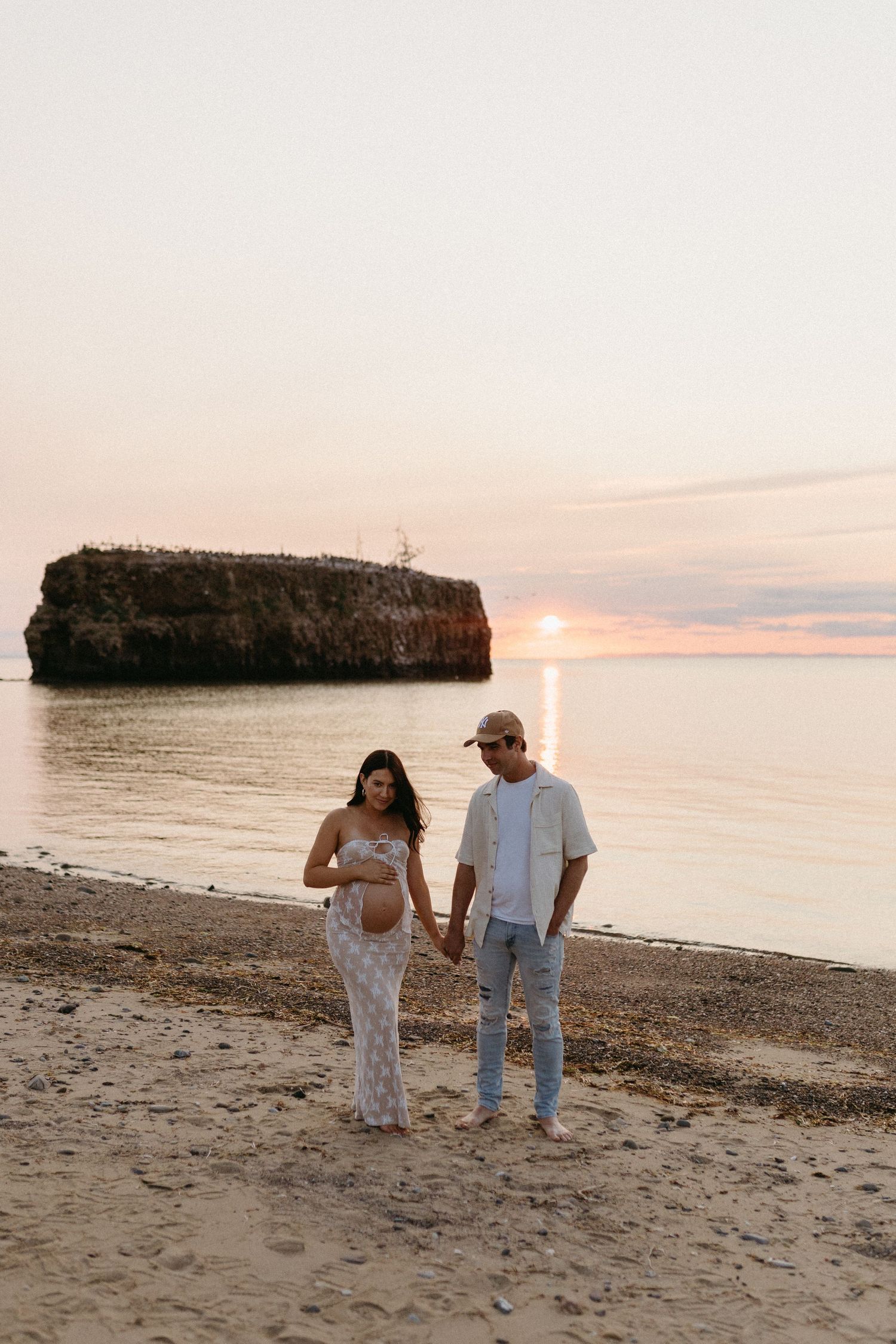 A couple strolls hand in hand along a peaceful beach at sunset with rocky cliffs in the background.