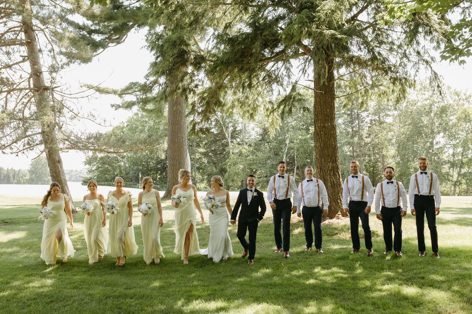 Wedding party stands in a row under tall trees in an outdoor garden setting during a summer celebration.