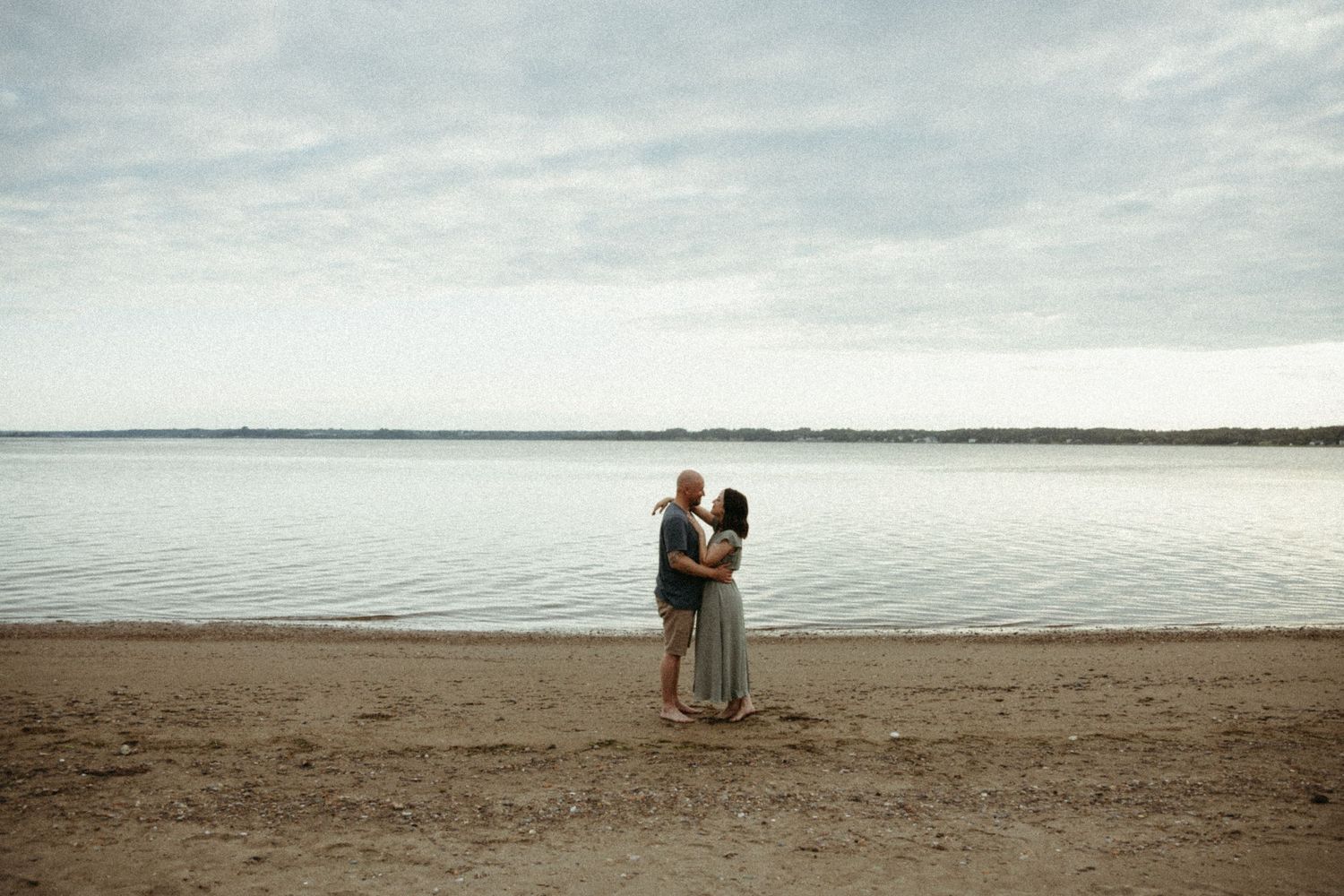 Couple embracing on a serene beach during sunset with calm ocean waters in the background.