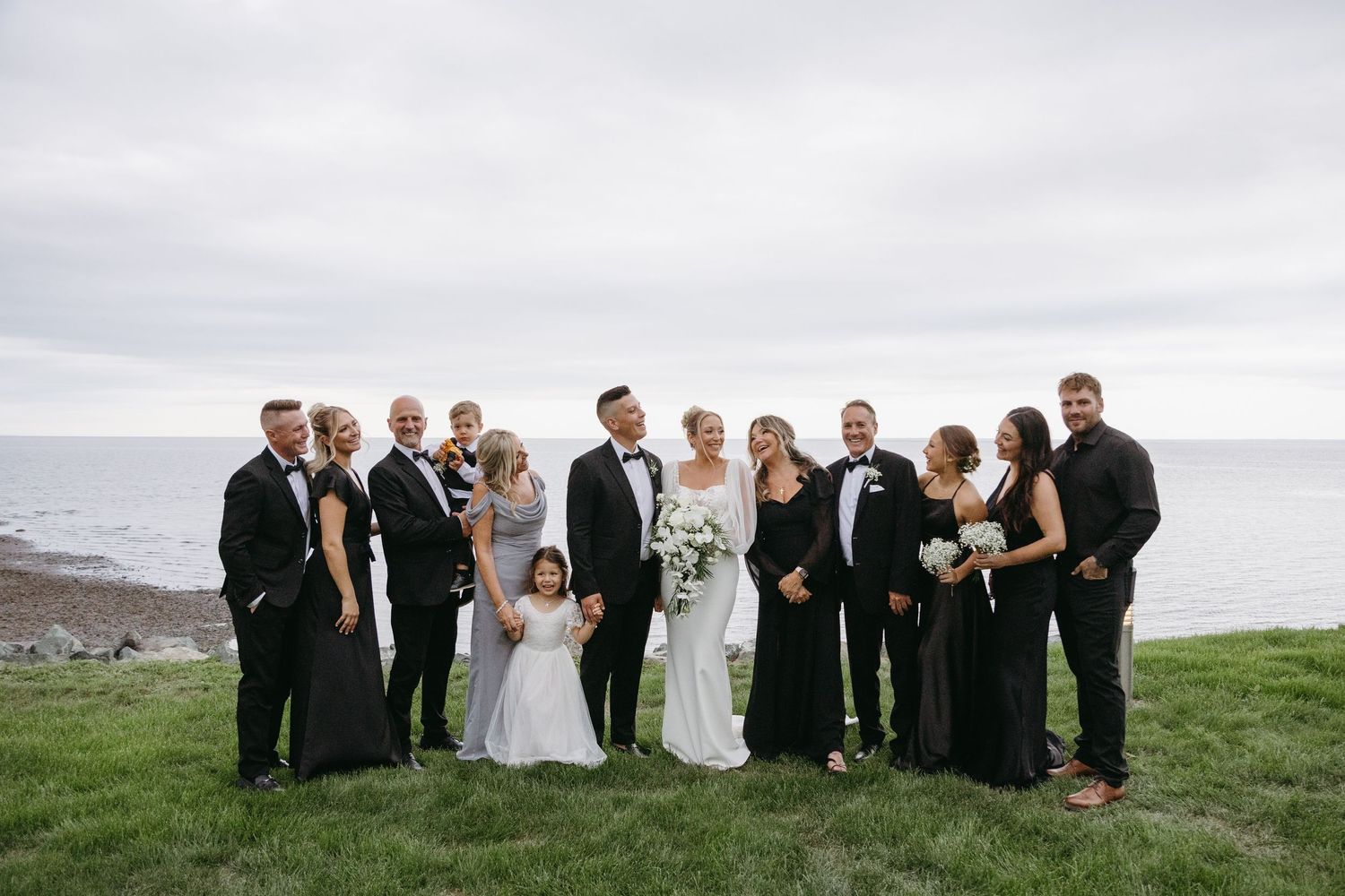 Wedding party in formal attire poses together on a scenic hillside overlooking a cloudy landscape.
