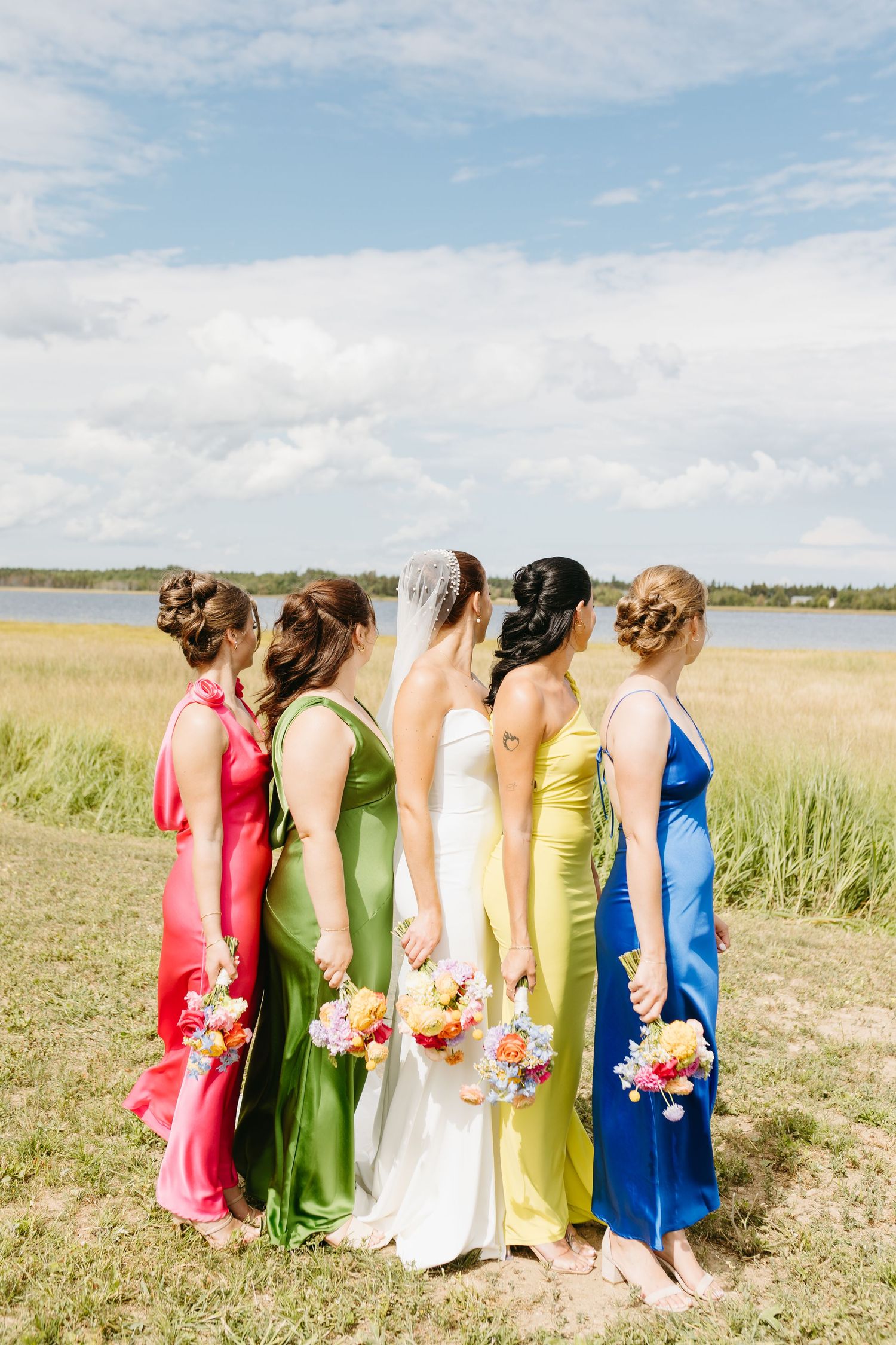 Group of bridesmaids in colorful rainbow dresses stand together in a grassy field by the water on a sunny day.