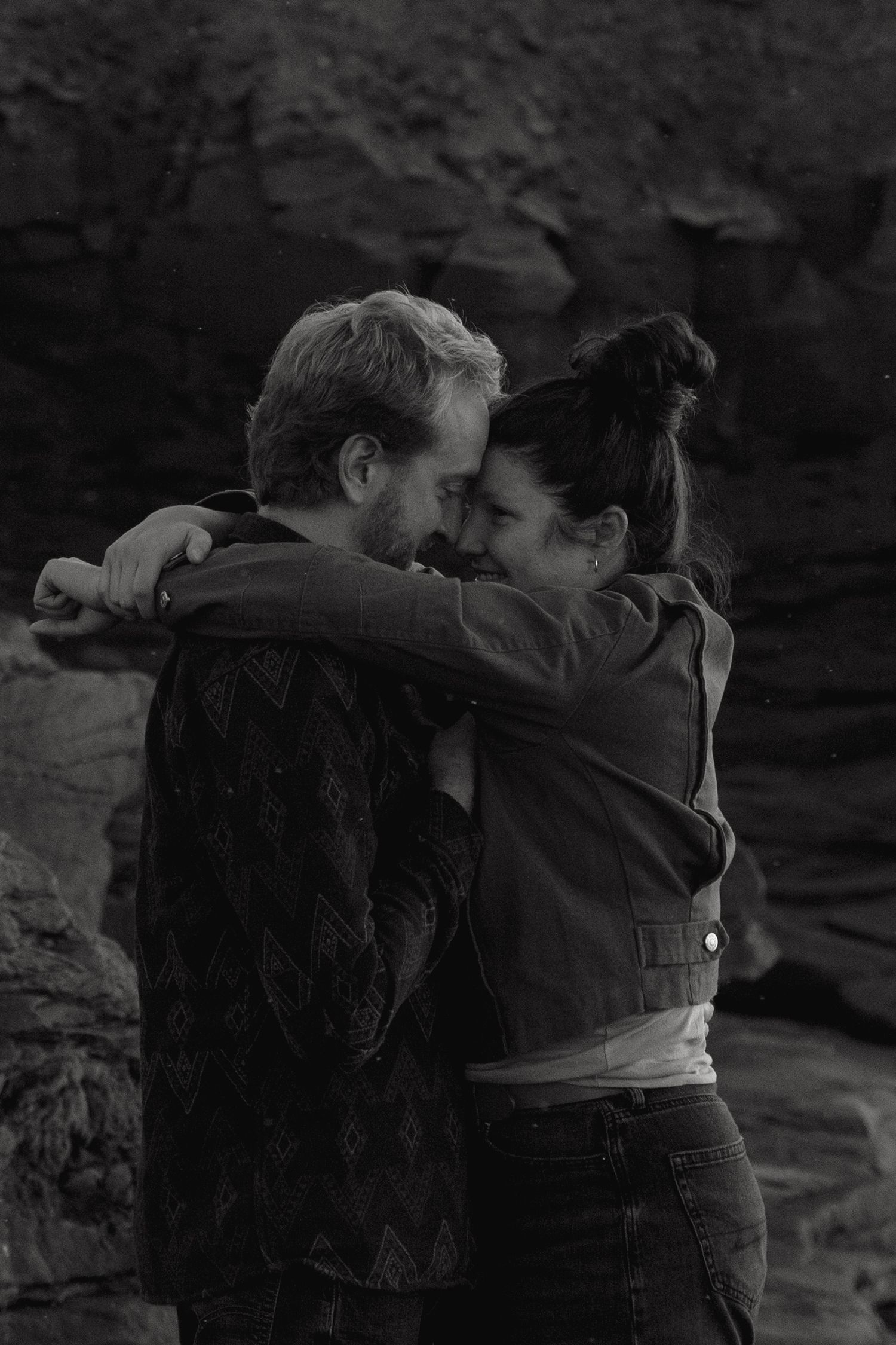 Two people embrace in a dark, dramatic black and white scene against a rocky backdrop.