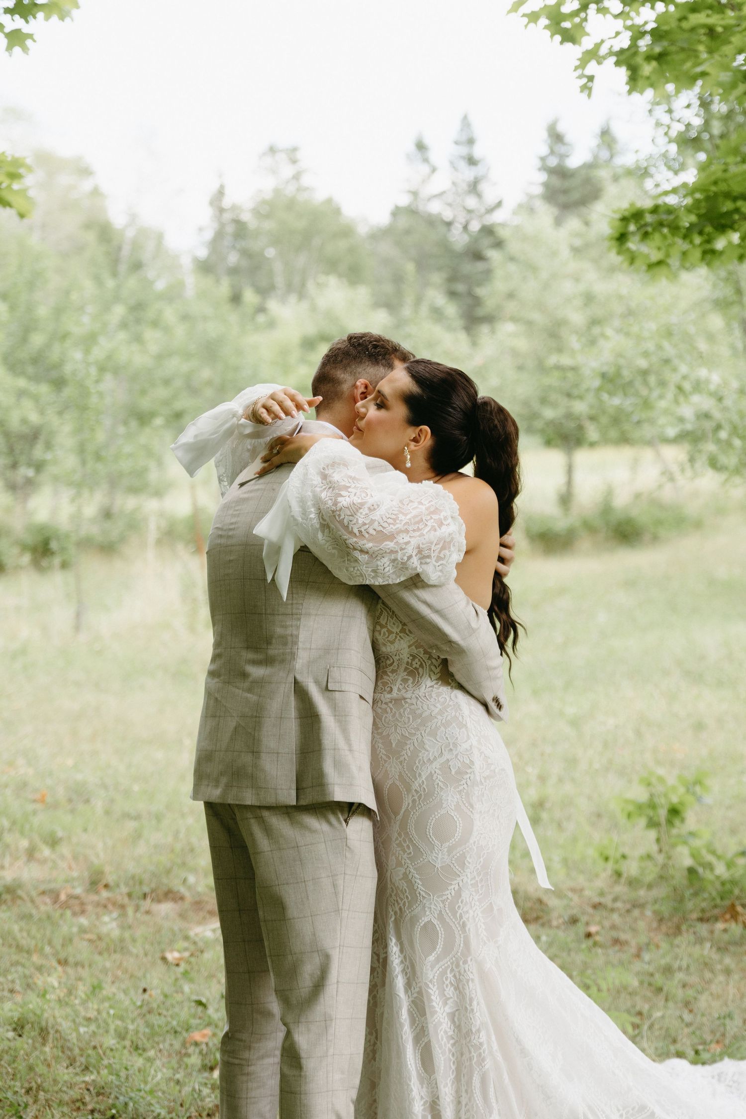 A couple shares an intimate moment in a sunlit meadow with lush greenery and trees in the background.