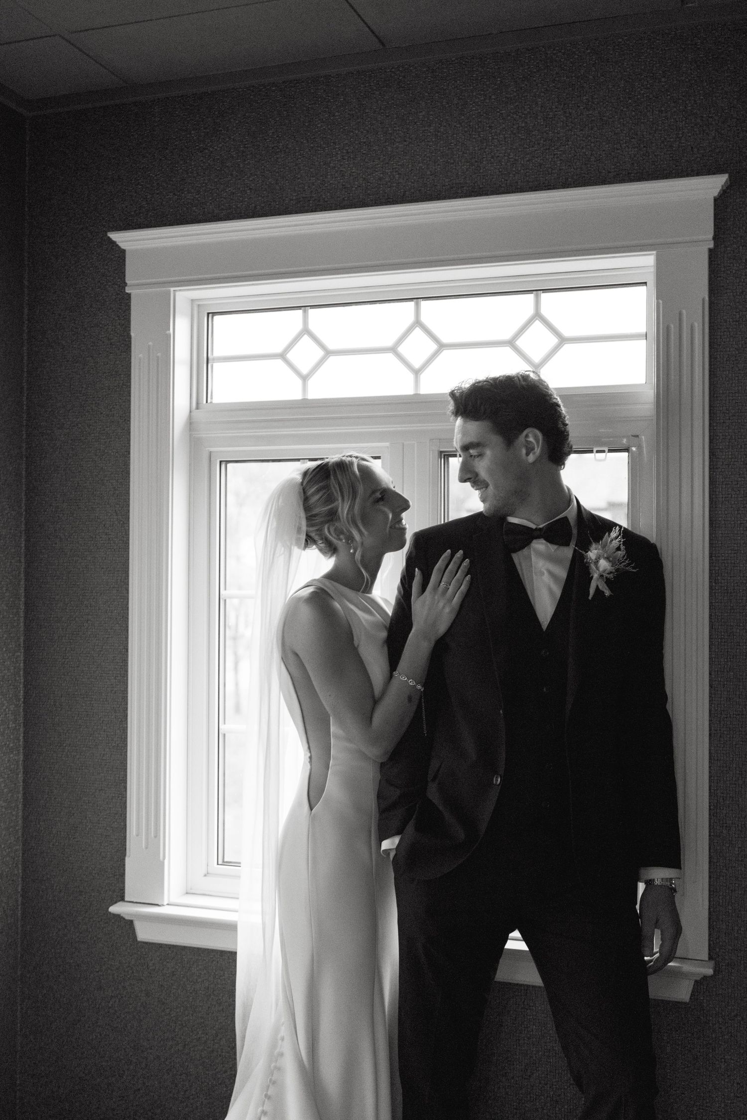 Black and white wedding photo of couple standing by window with decorative glass panes in elegant formal attire.