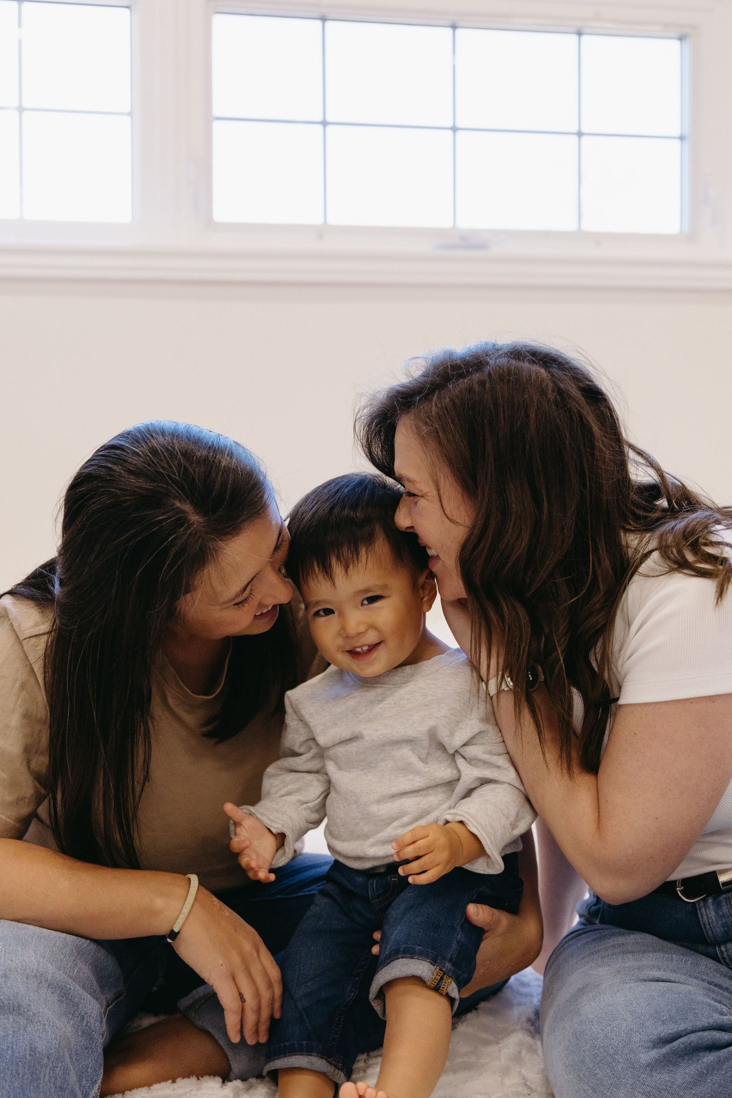 A heartwarming family moment shows three people sharing affectionate smiles while sitting together on a couch by a window.
