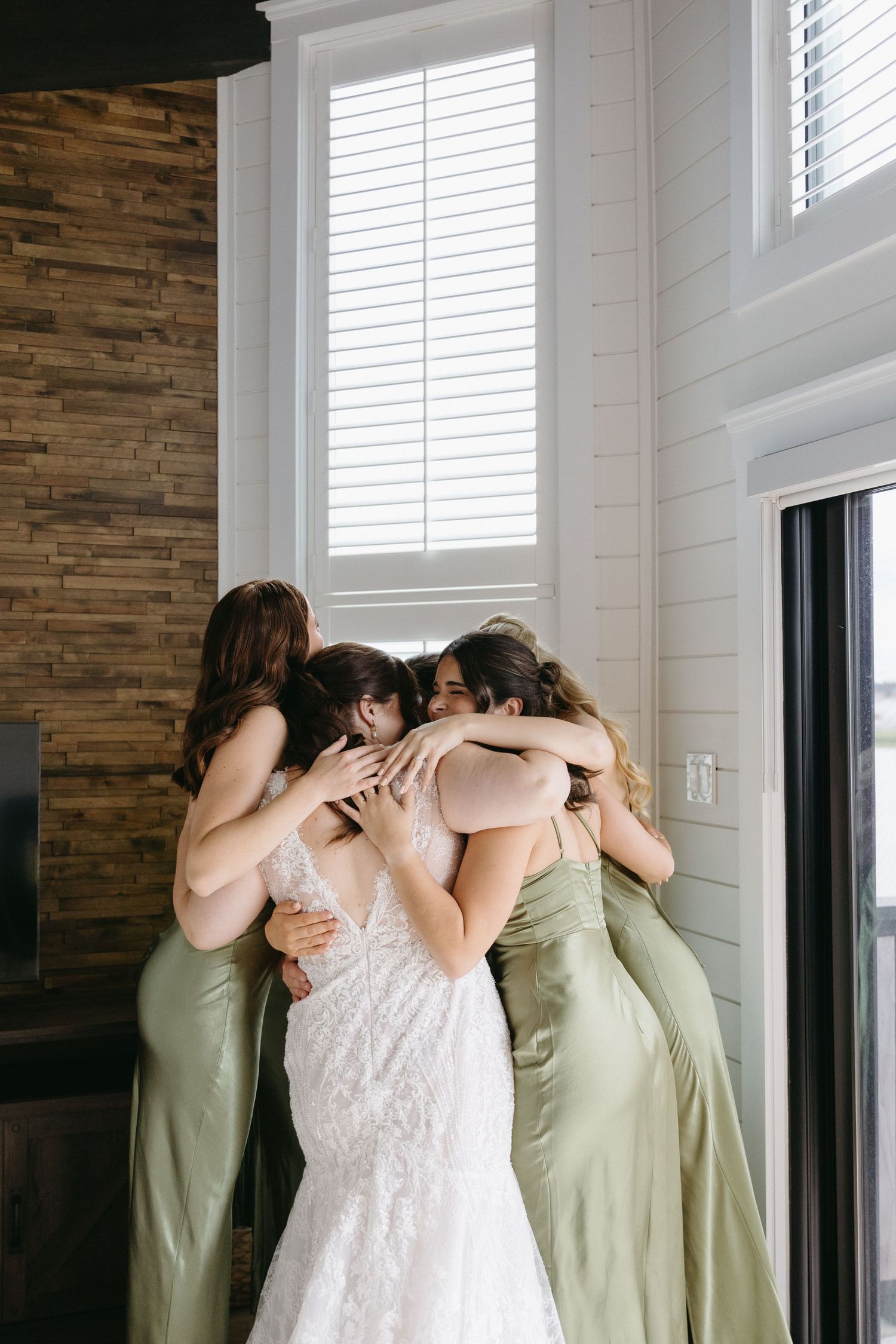 A group in sage green dresses and white lace gather in an emotional huddle by a bright window.