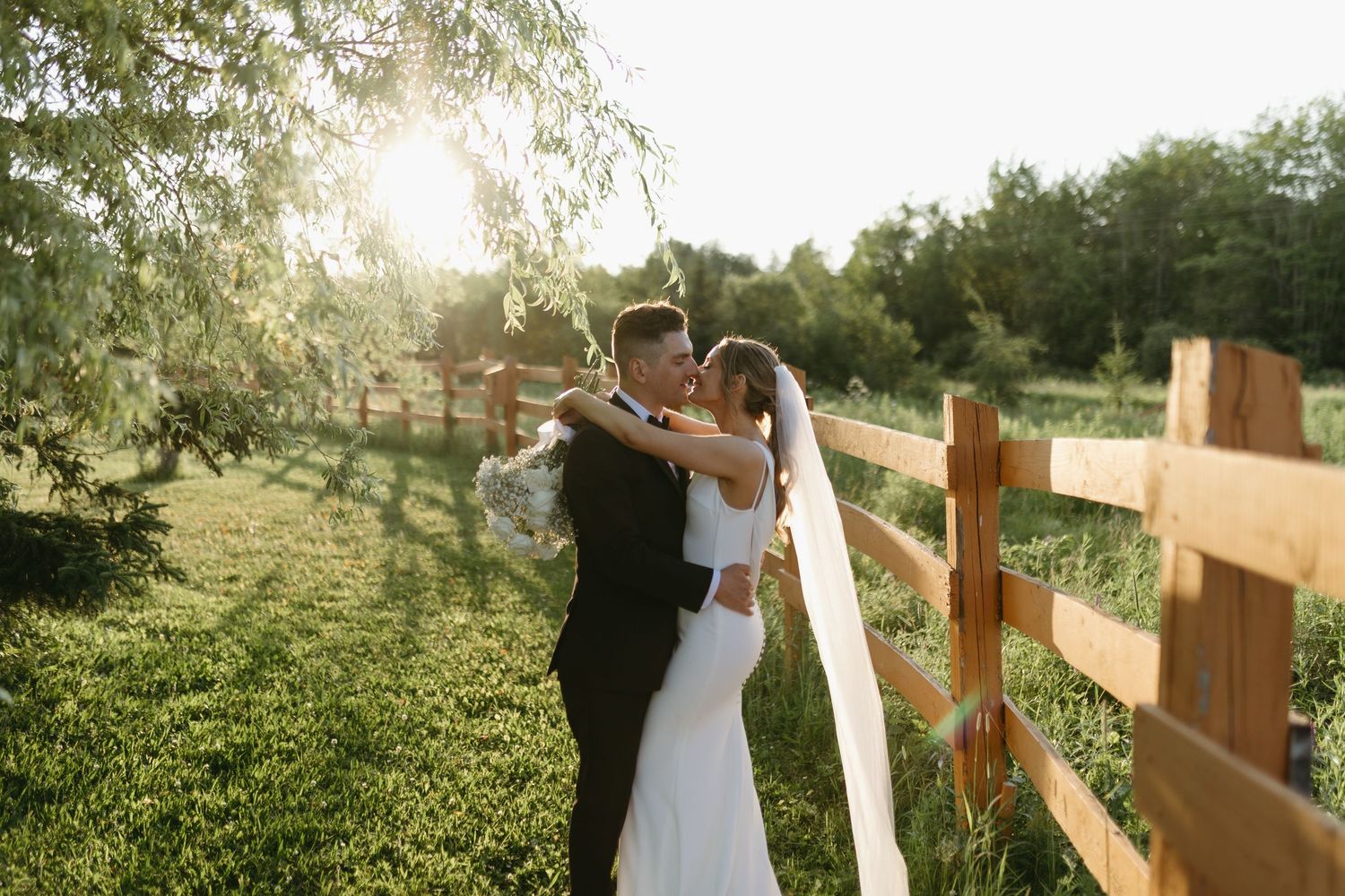 A couple shares a romantic moment at sunset by a wooden fence in a rustic outdoor setting.