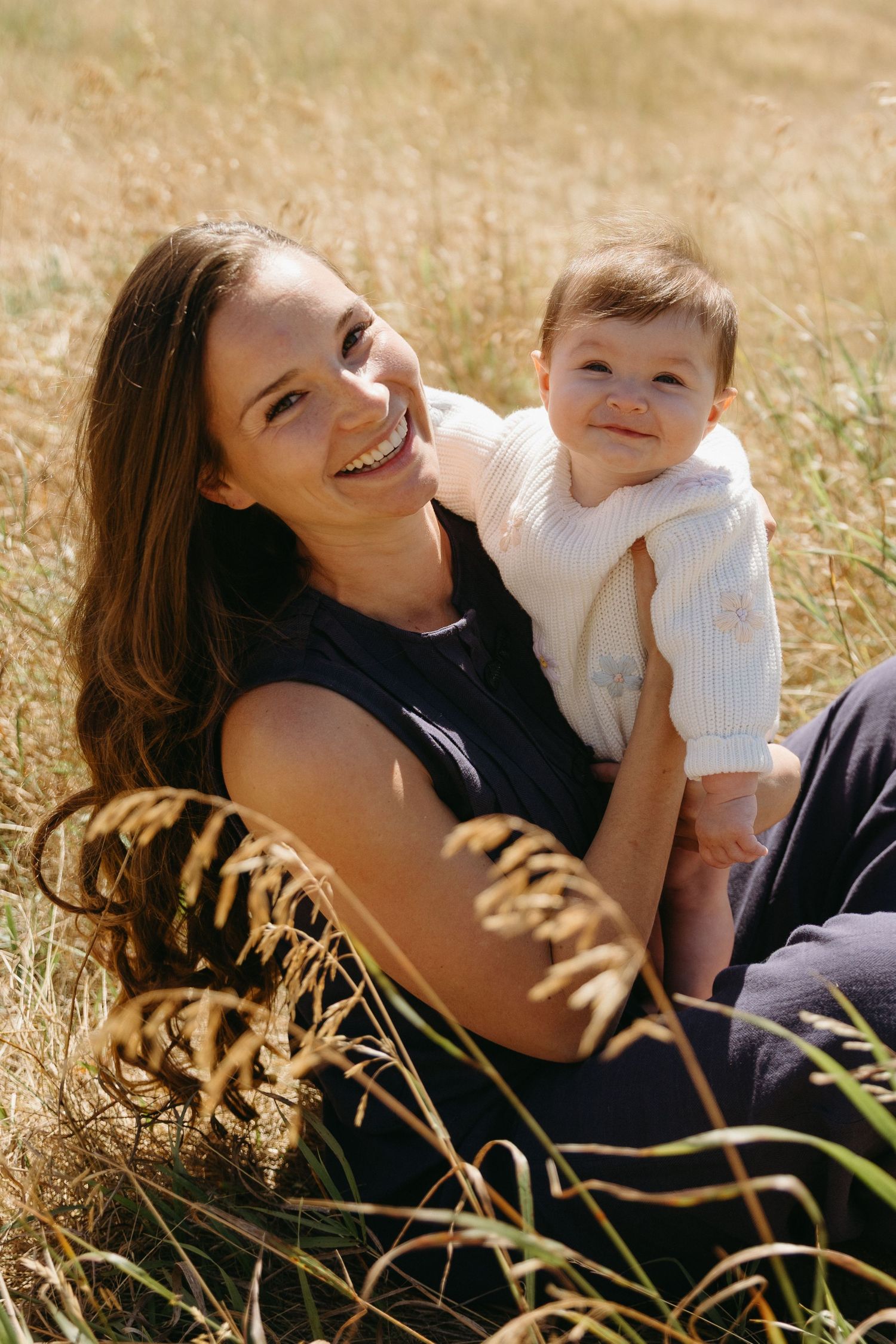 A parent and baby sit together in a sunlit field of tall golden grass on a warm afternoon.