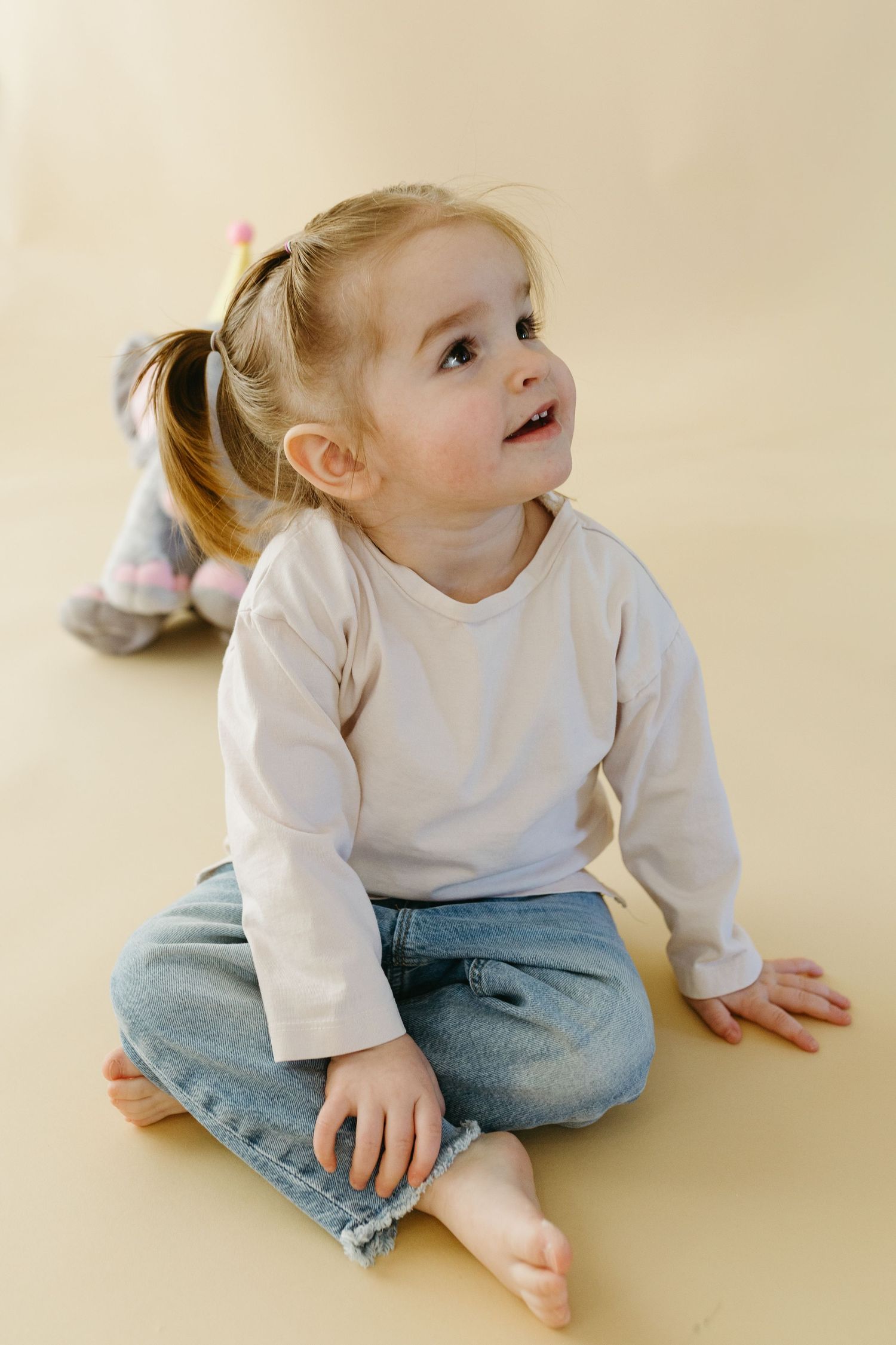 A toddler in a white long sleeve shirt and jeans sits on a beige floor looking upward with wonder.