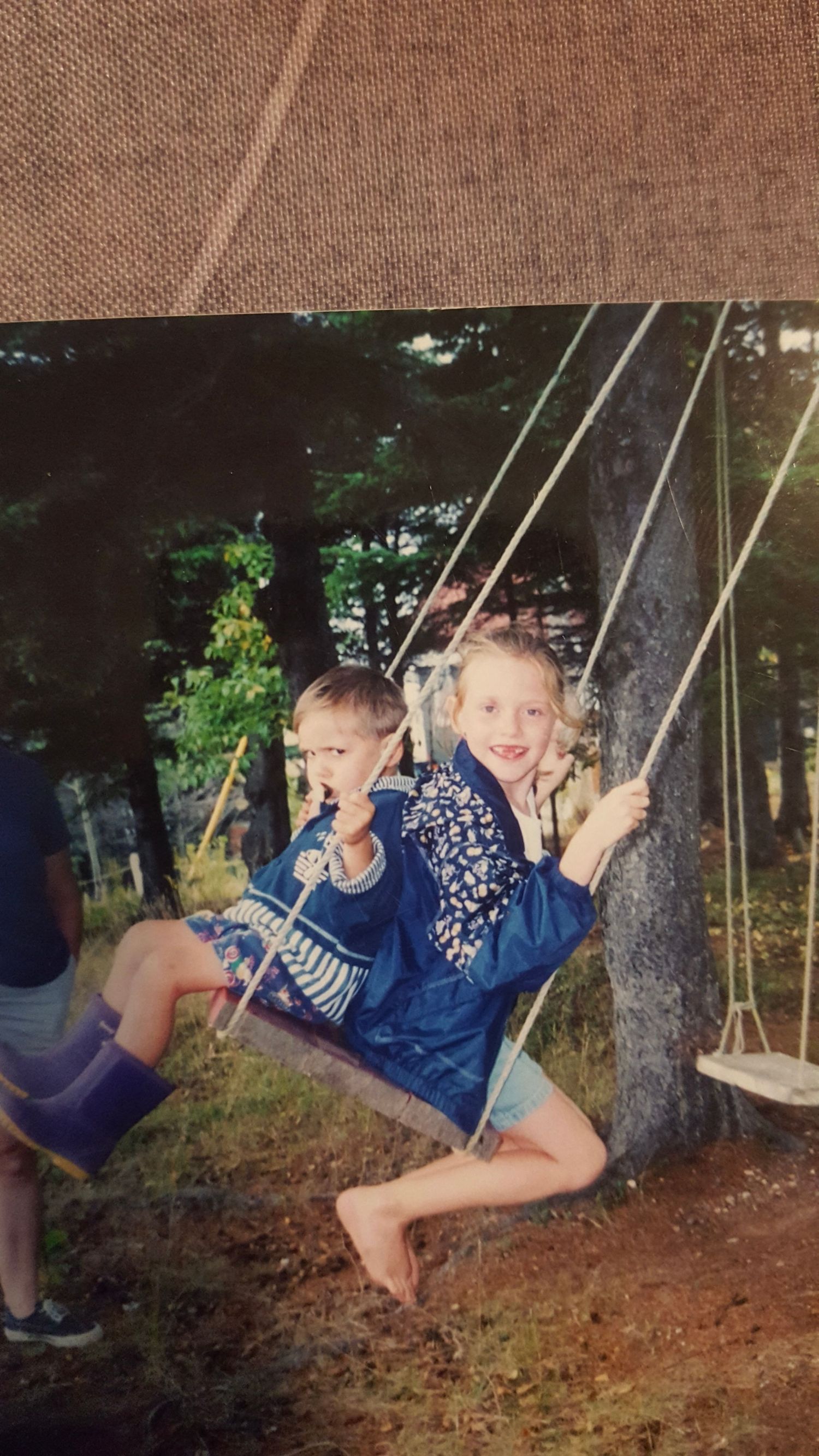 Two children in blue outfits enjoy swinging together on a backyard rope swing at dusk.