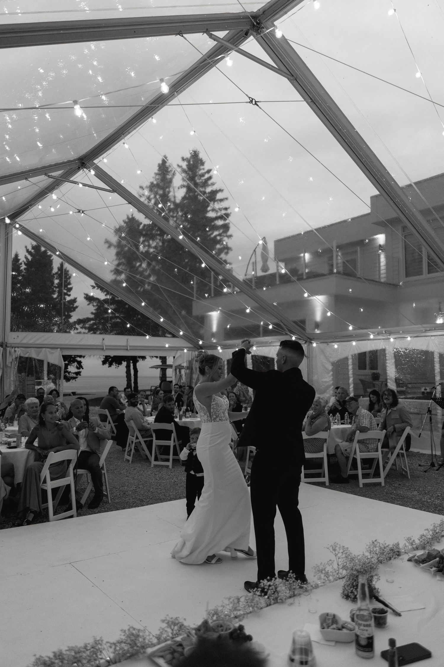 Wedding reception guests dance and celebrate under a white tent decorated with twinkling lights and floral arrangements.
