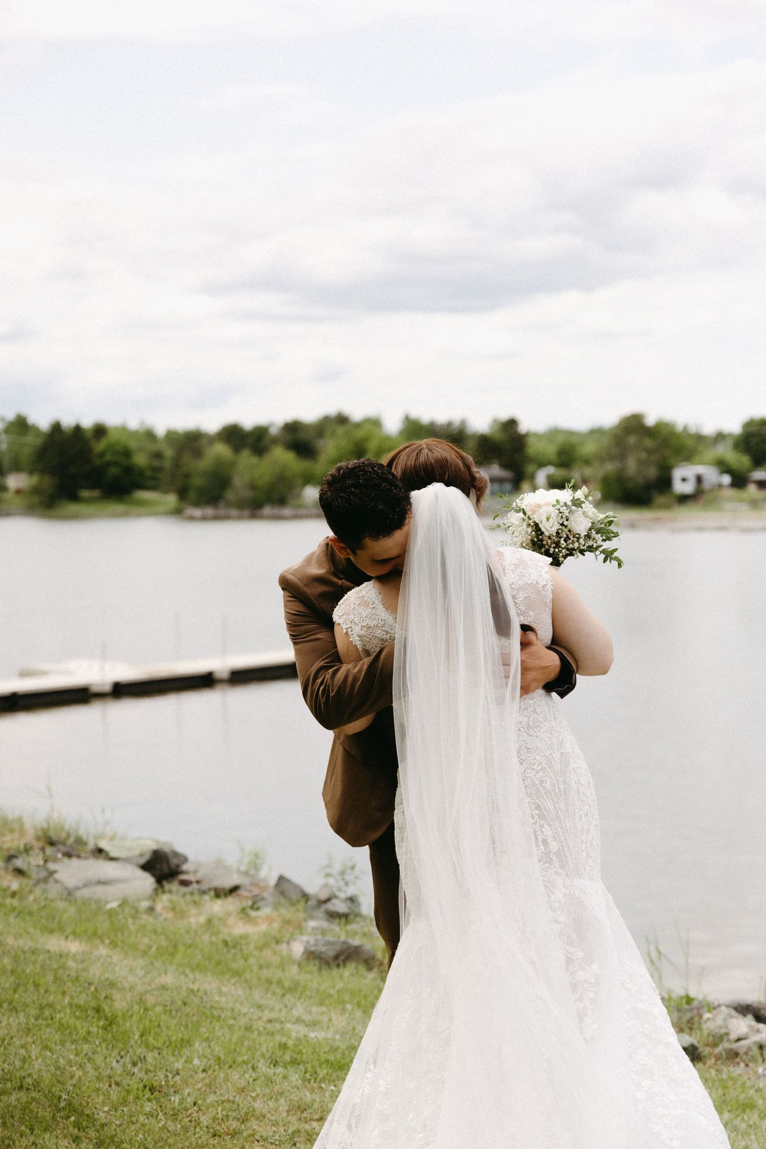 A flowing white wedding veil blows in the wind against a scenic waterfront backdrop on a cloudy day.
