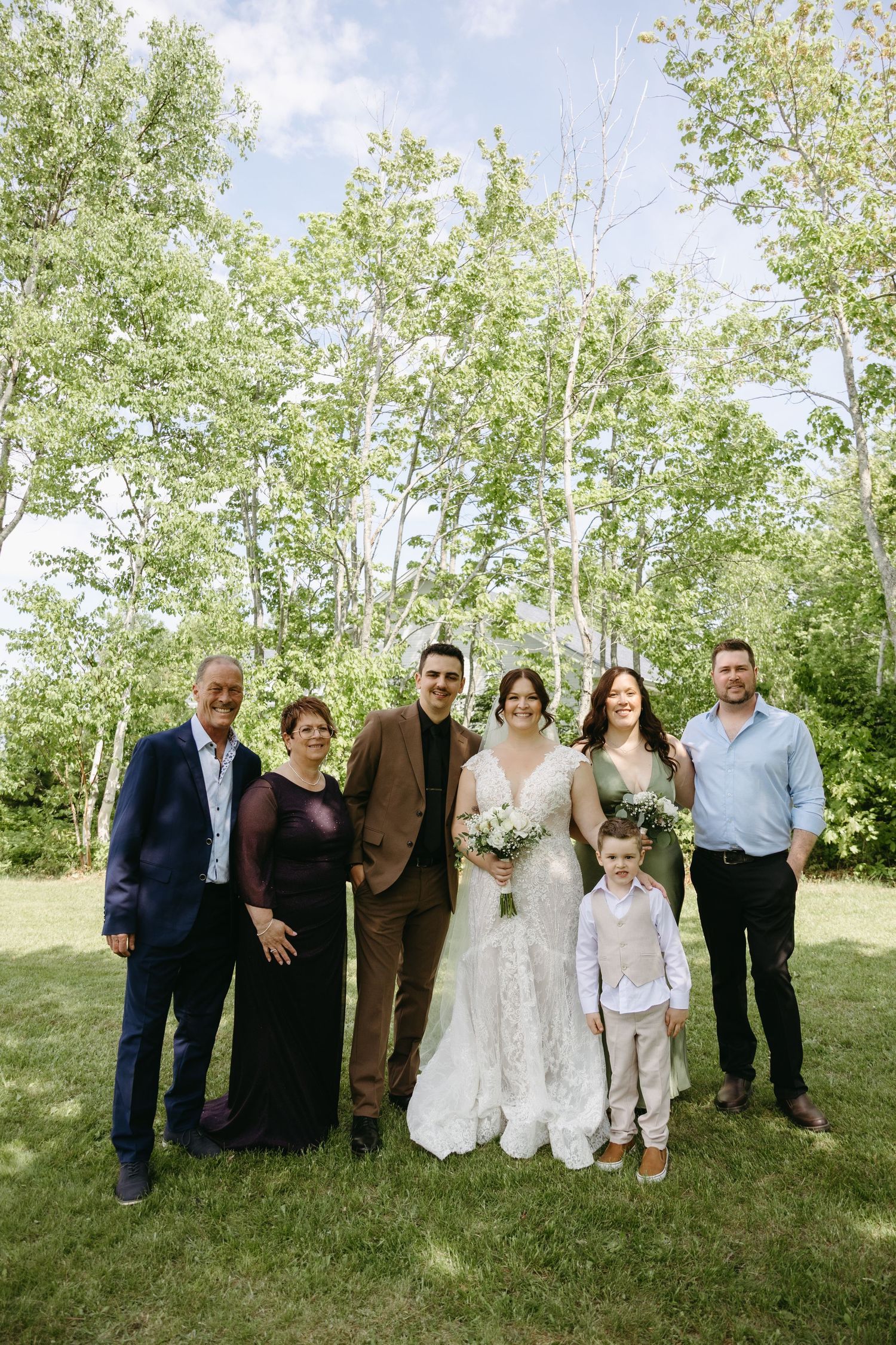 A wedding party stands together on a grassy lawn with trees in the background during an outdoor celebration.