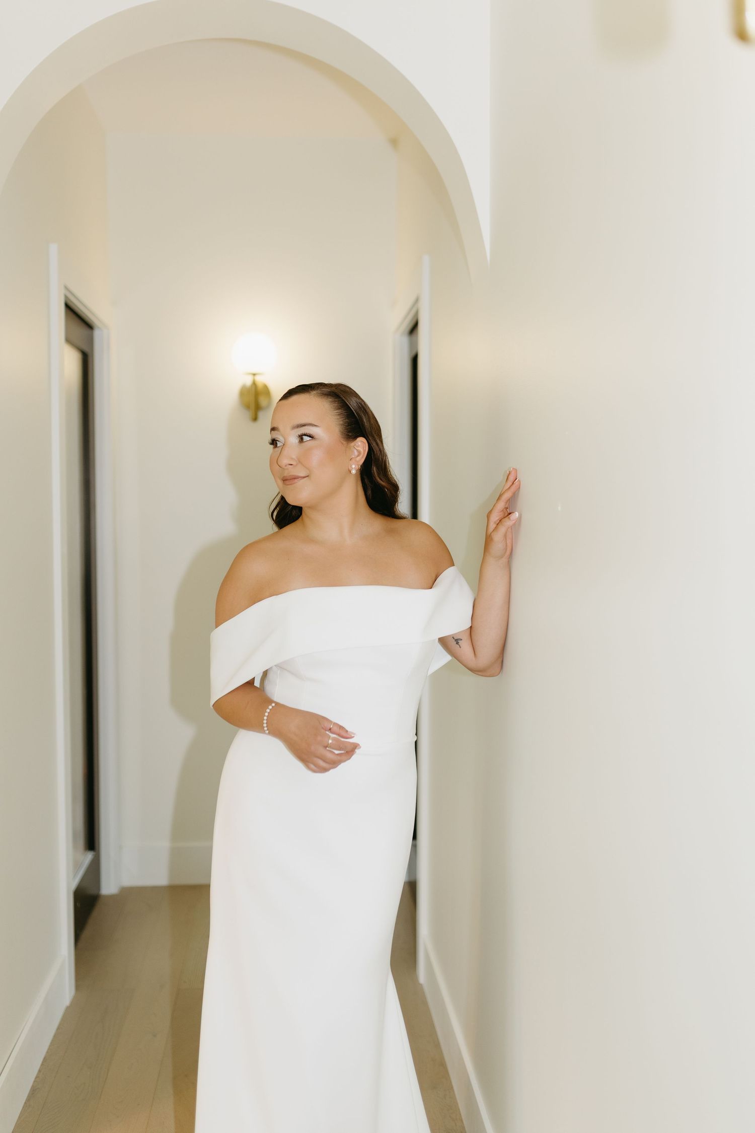 A person wrapped in a white towel poses against a white wall in a minimalist hallway setting.