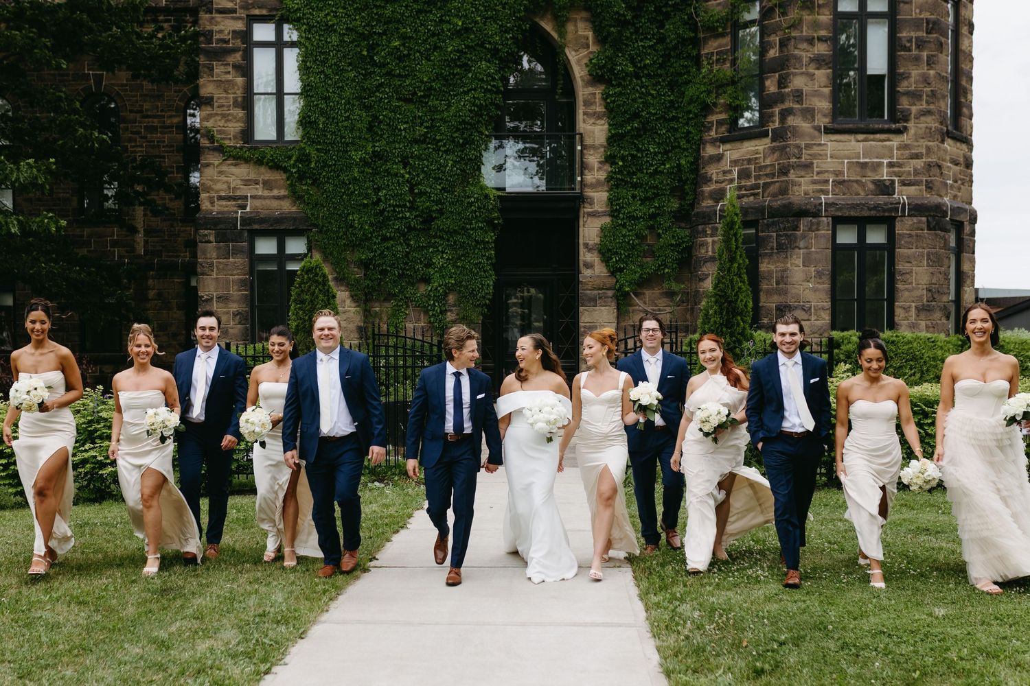 Wedding party poses in a line against ivy-covered stone building exterior in elegant formal attire.