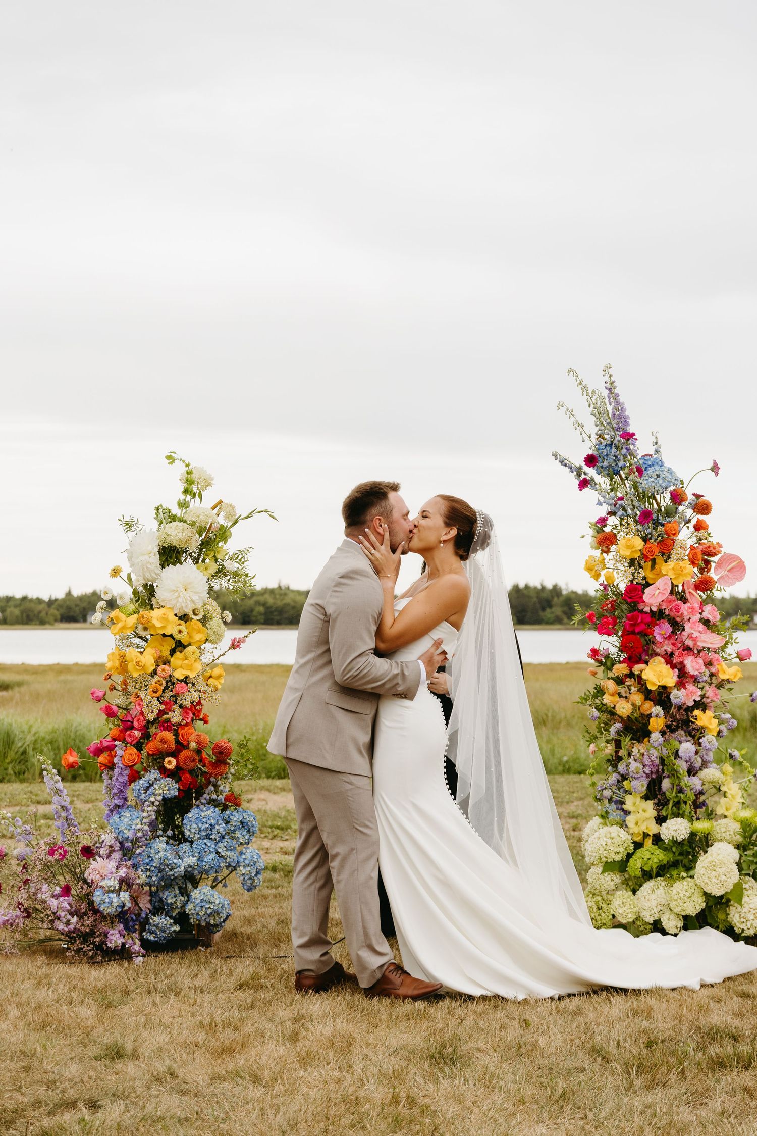 A wedding couple shares a kiss outdoors between two vibrant floral arrangements with colorful blooms by a lake.