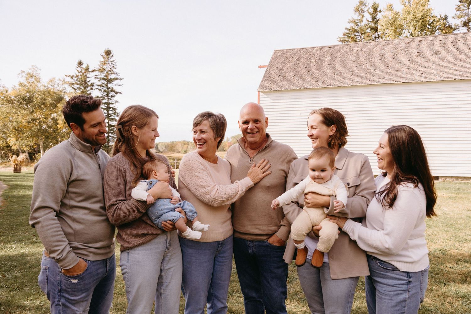 A group in neutral colored sweaters laughs together while standing outside a white barn on a sunny autumn day.