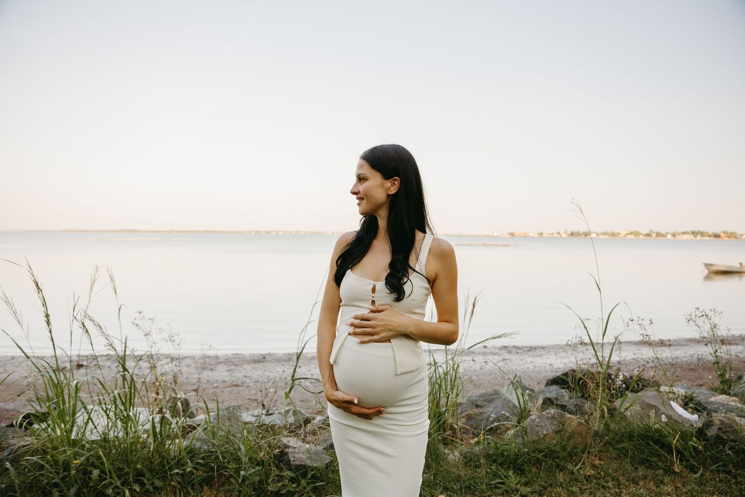 A pregnant woman in a white dress poses in a grassy field during golden hour for a maternity photoshoot.