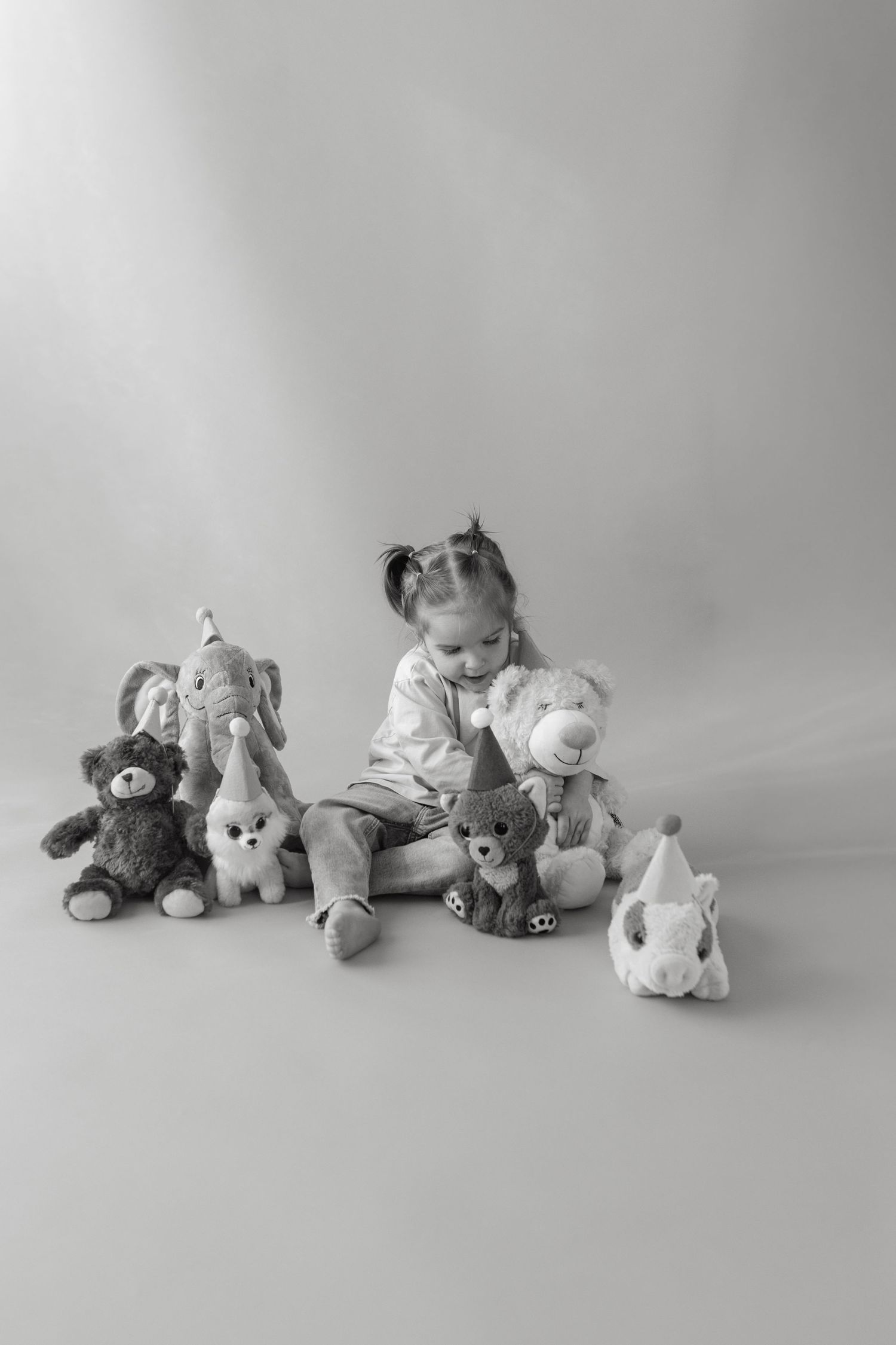 A black and white photo of a child sitting with a collection of stuffed animals against a plain backdrop.