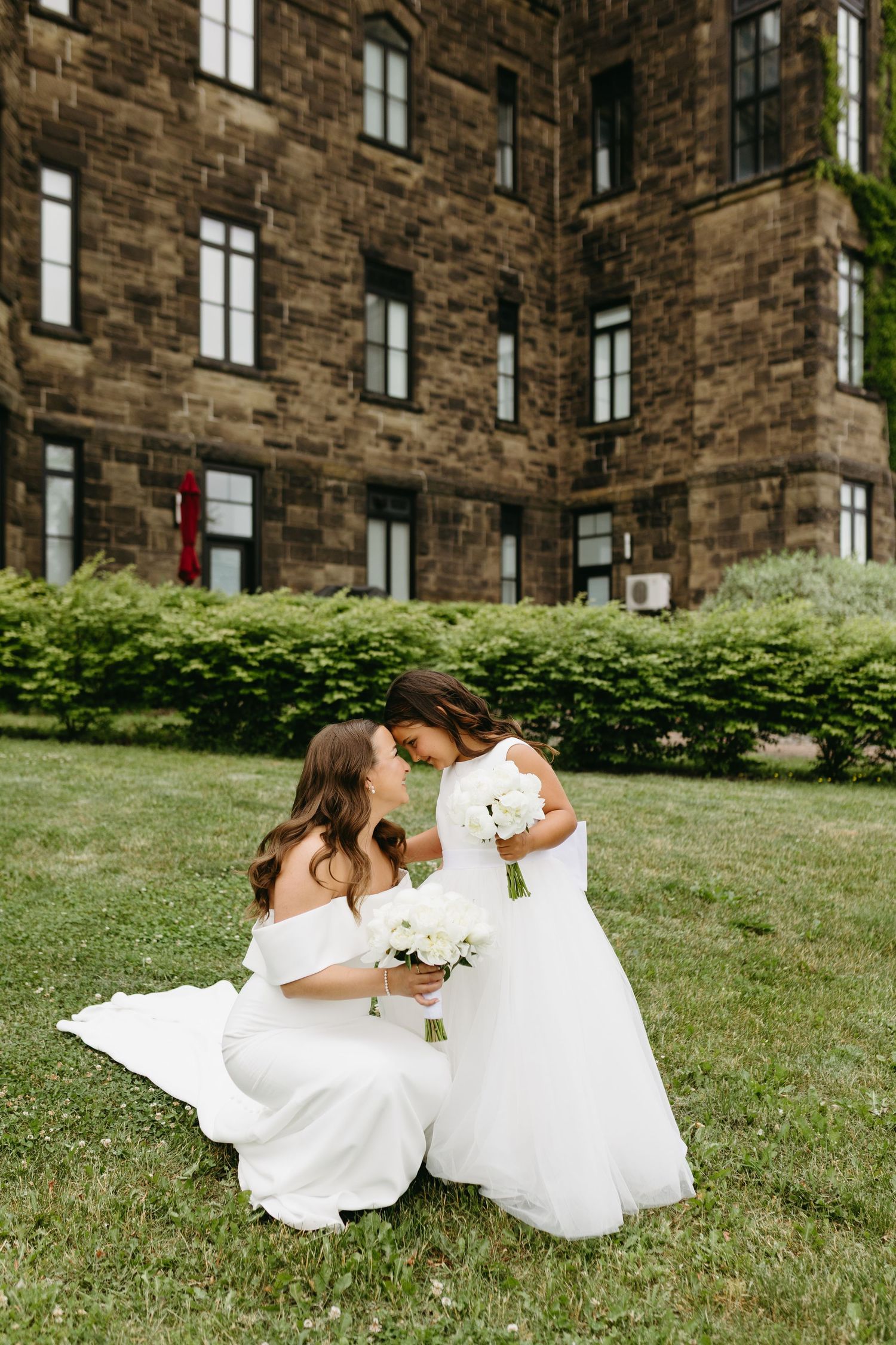 Two people in white wedding dresses lay together on a grassy hill beside a brick building.