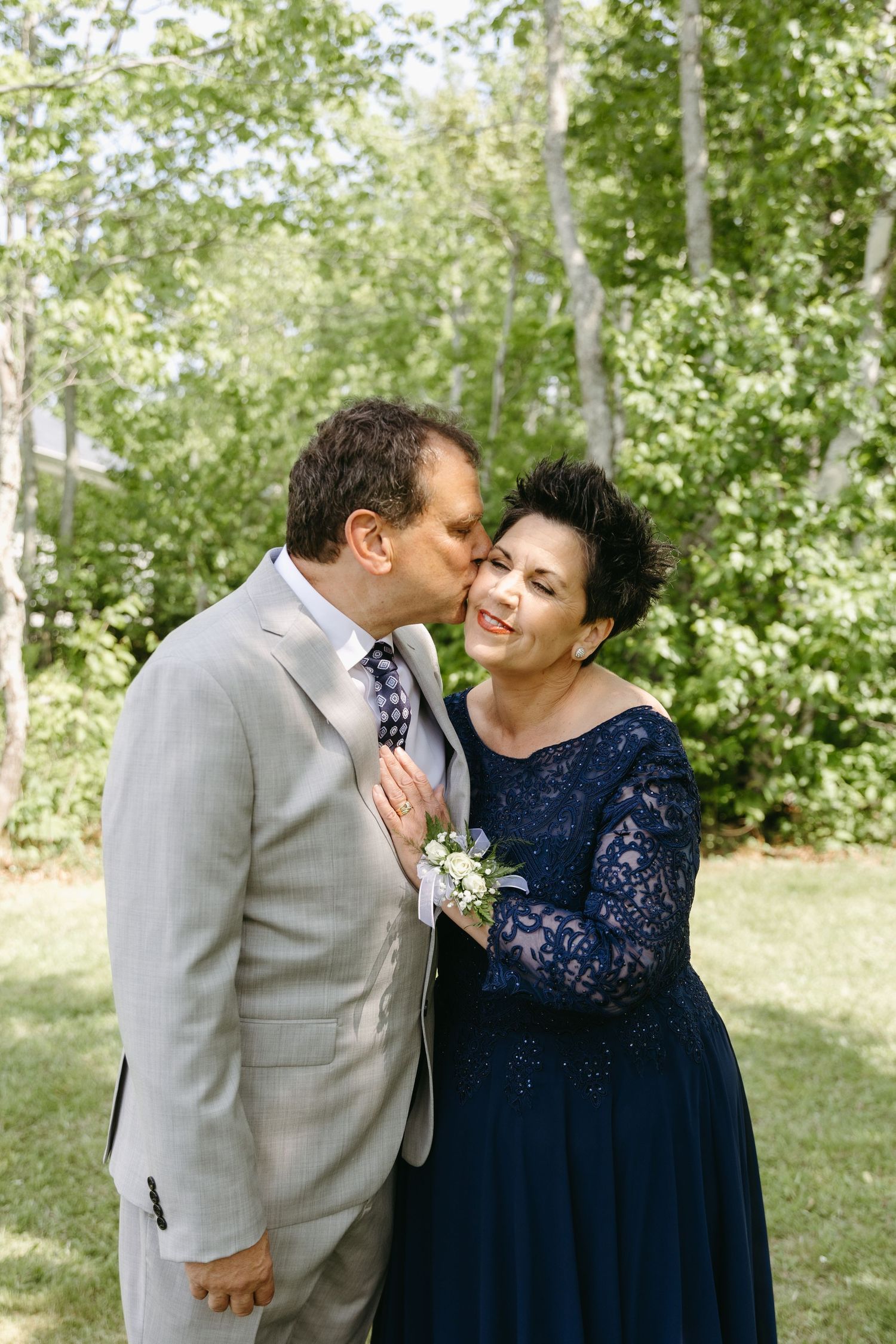 A tender moment captured between two people in a navy dress and gray suit standing outdoors among green trees.