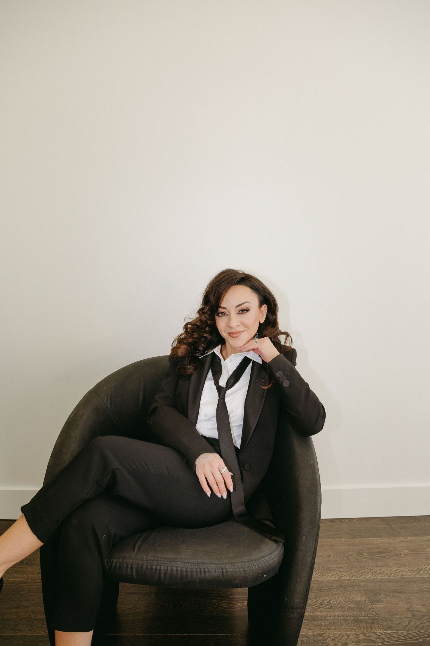 Professional portrait of someone in a black suit and white tie lounging in a dark armchair against a light wall.