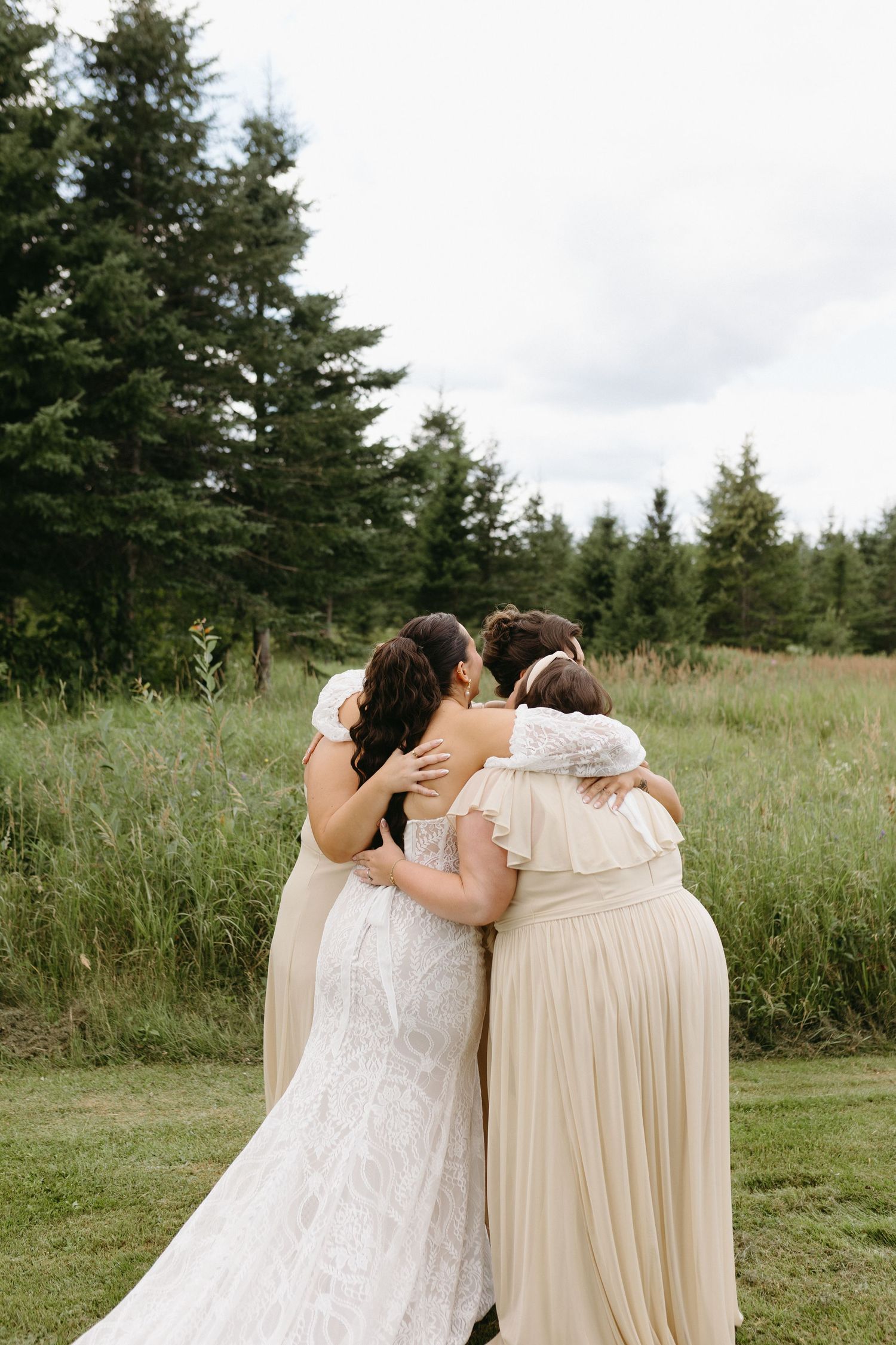 Three people embrace while standing in a grassy meadow surrounded by evergreen trees on an overcast day.