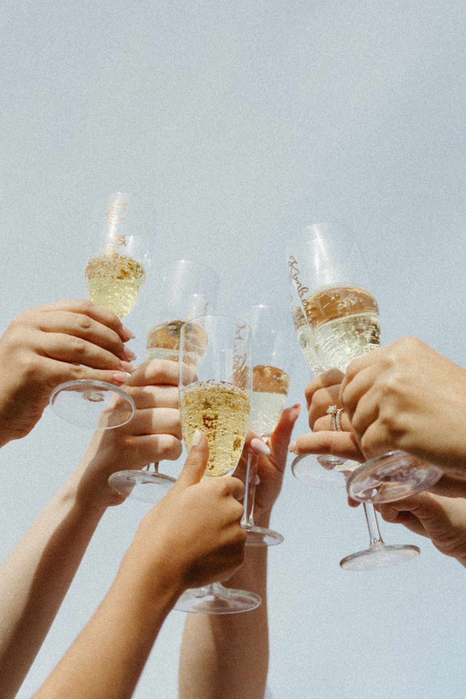 Multiple hands raise champagne glasses in a celebratory toast against a light gray background.