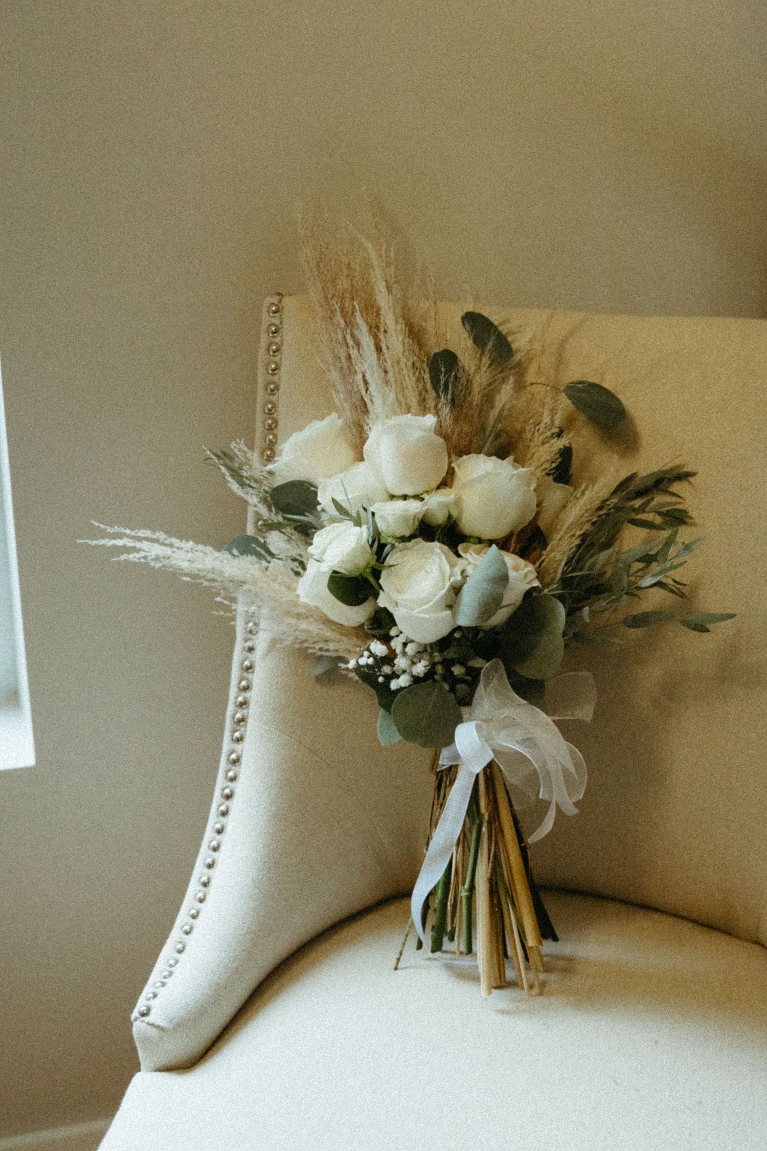 A white floral bouquet with dried elements rests against a beige cushioned surface, showing multiple angles.