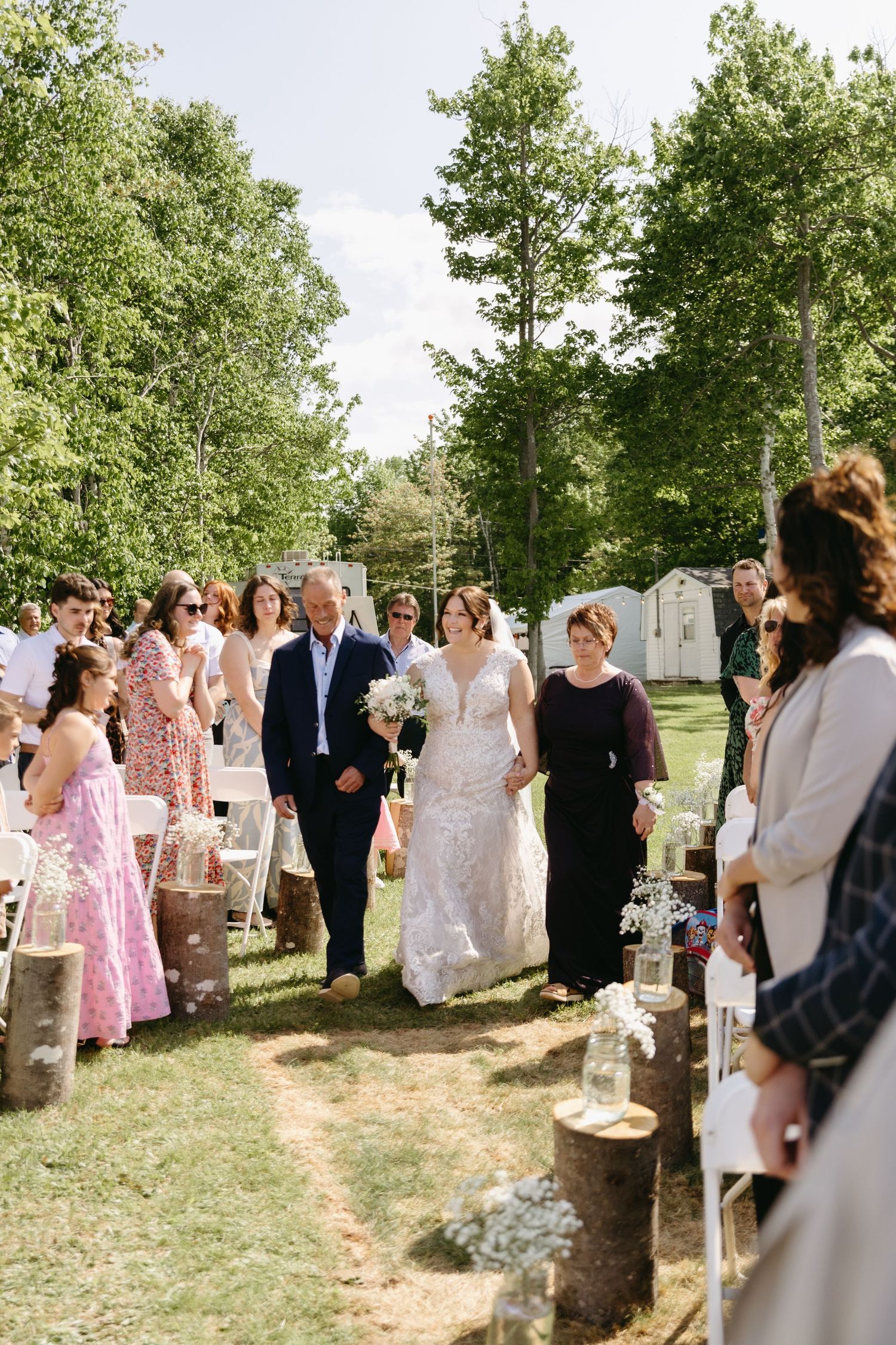 Wedding party and guests gather outdoors under trees for a summer celebration in natural setting.