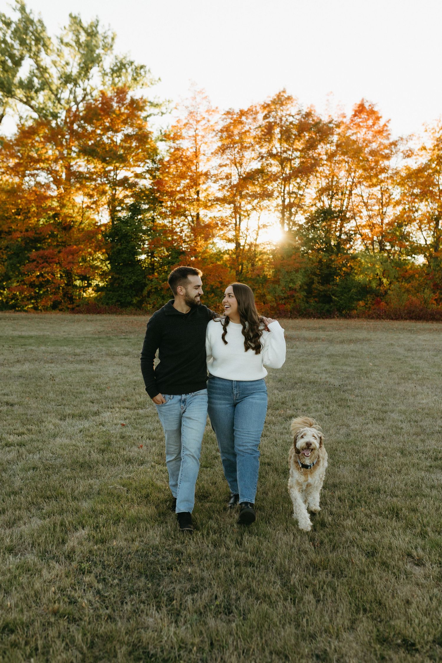 A couple walks with their fluffy golden doodle dog through a grassy field at sunset during autumn.