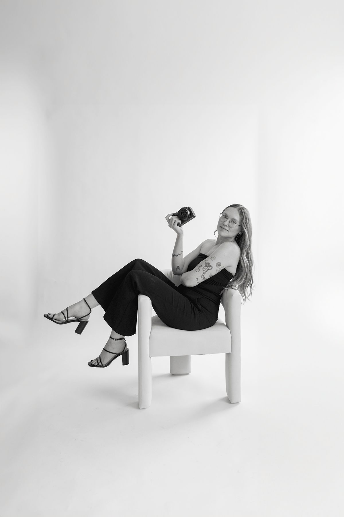 A woman in black attire lounges casually in a white chair against a white backdrop while holding a camera.