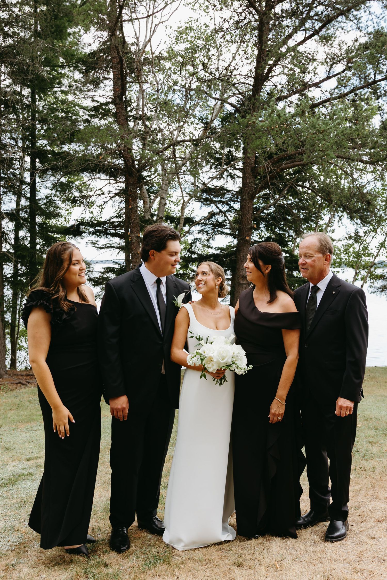 Wedding party in black and white attire laughing together in a casual candid sequence outdoors near pine trees.