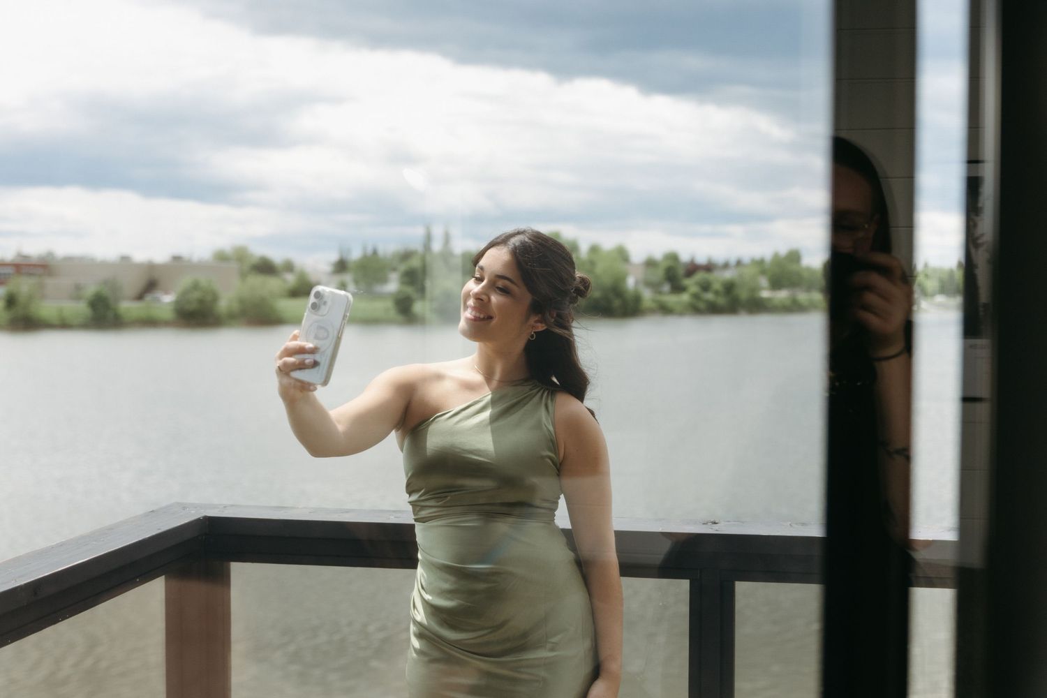 Someone in a green dress takes a selfie while standing on a lakeside deck on a cloudy day.