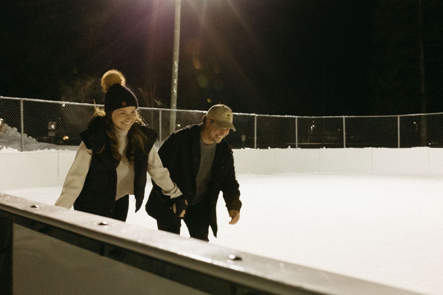Two people ice skating at night on an outdoor rink illuminated by overhead lights in winter.